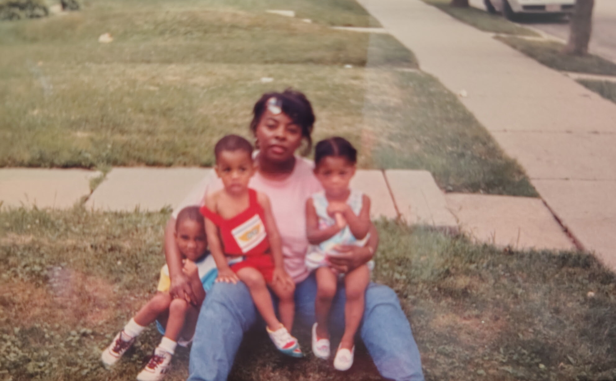 A woman sits on the grass outdoors with four young children in a suburban yard, posing for a photo.