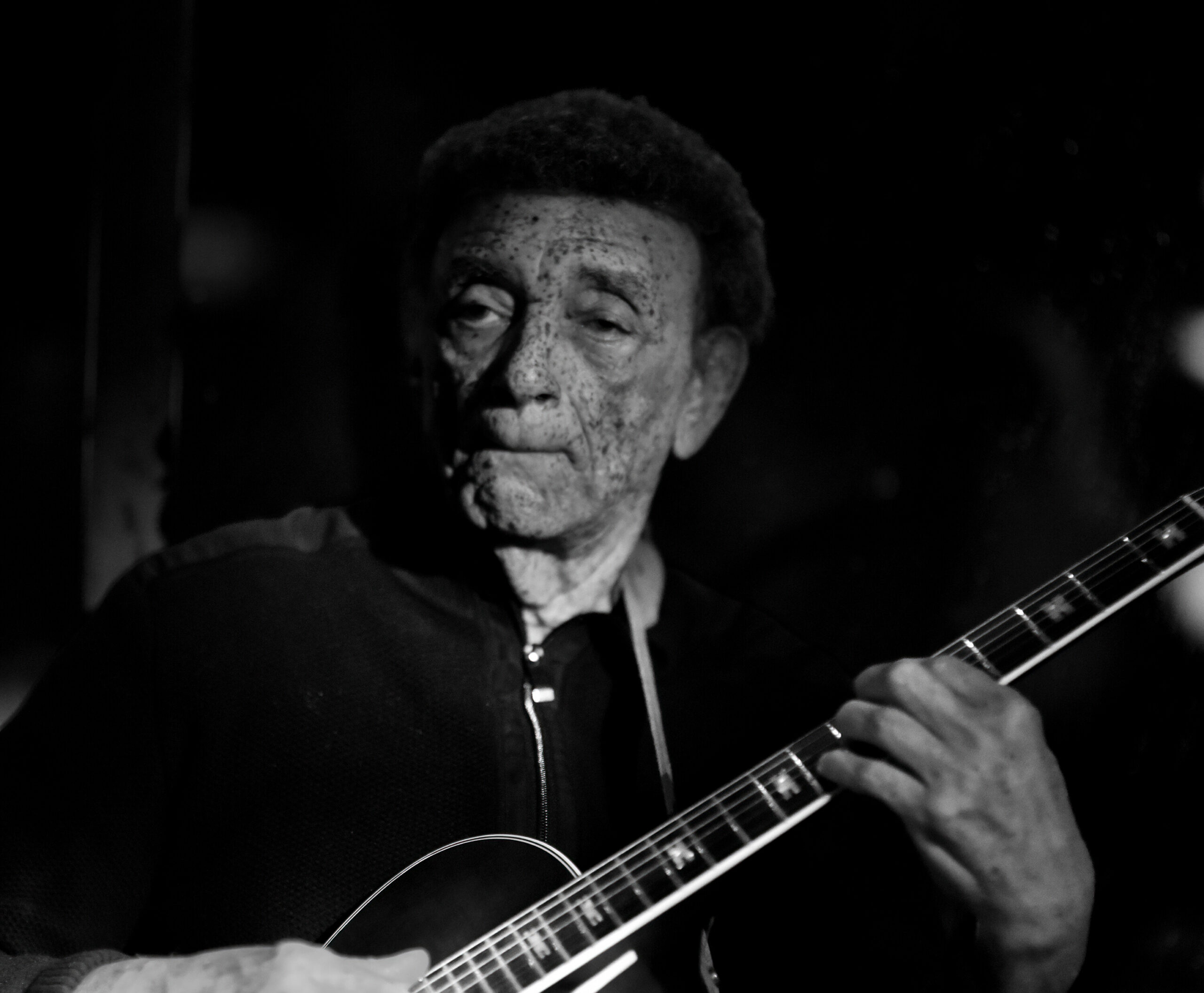 Close-up, black-and-white shot of an older man playing an acoustic guitar, with focus on his freckled face and hand on the fretboard.