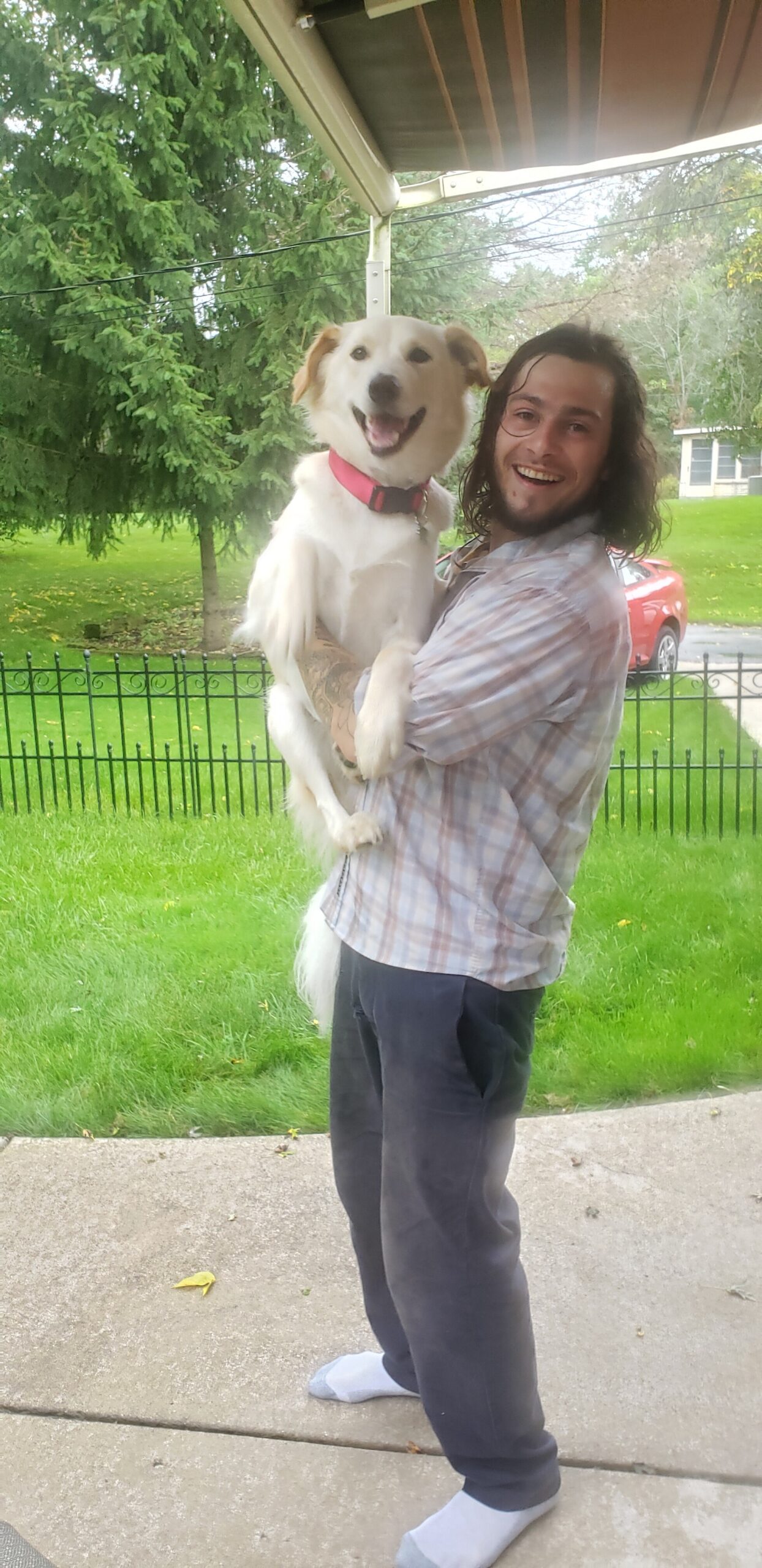 Man with shoulder-length hair and a plaid shirt smiles while holding a happy white dog with a pink collar on a porch.