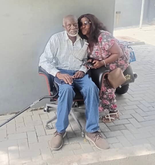 An elderly man sits in a wheelchair beside a woman who is leaning in and smiling outdoors on a sidewalk.