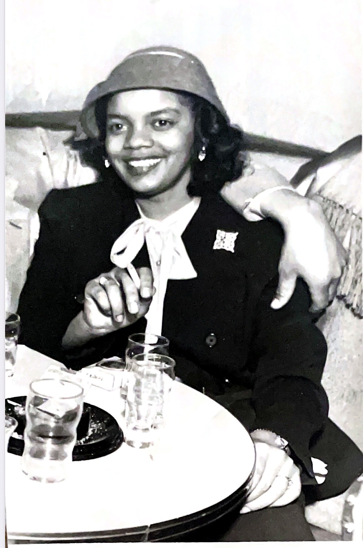 Smiling woman in a hat and blazer sits at a table with drinks, gesturing with her hand.