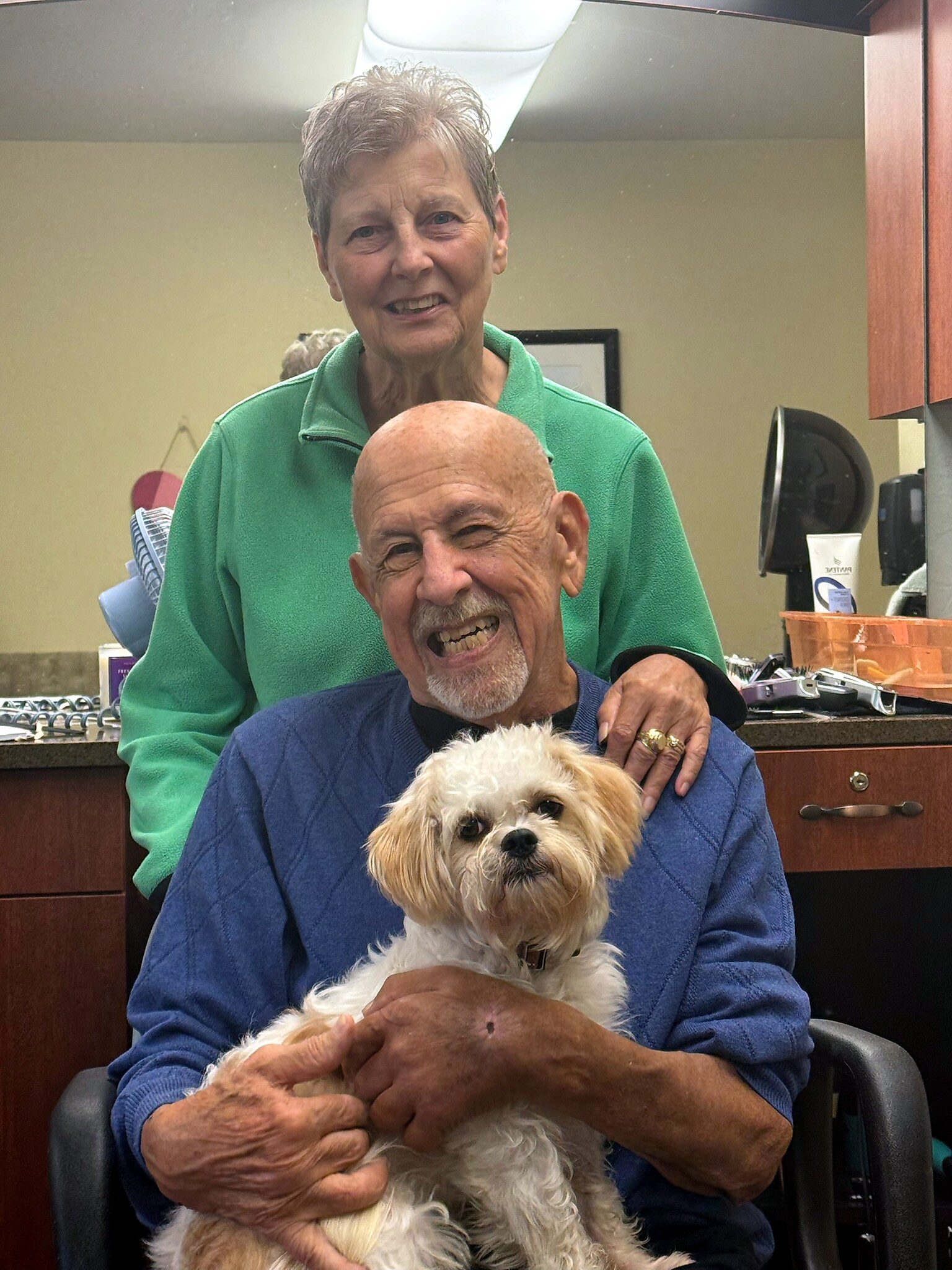 Smiling elderly man in a blue sweater sits in a salon chair holding a small fluffy dog on his lap, with a woman in green behind them.