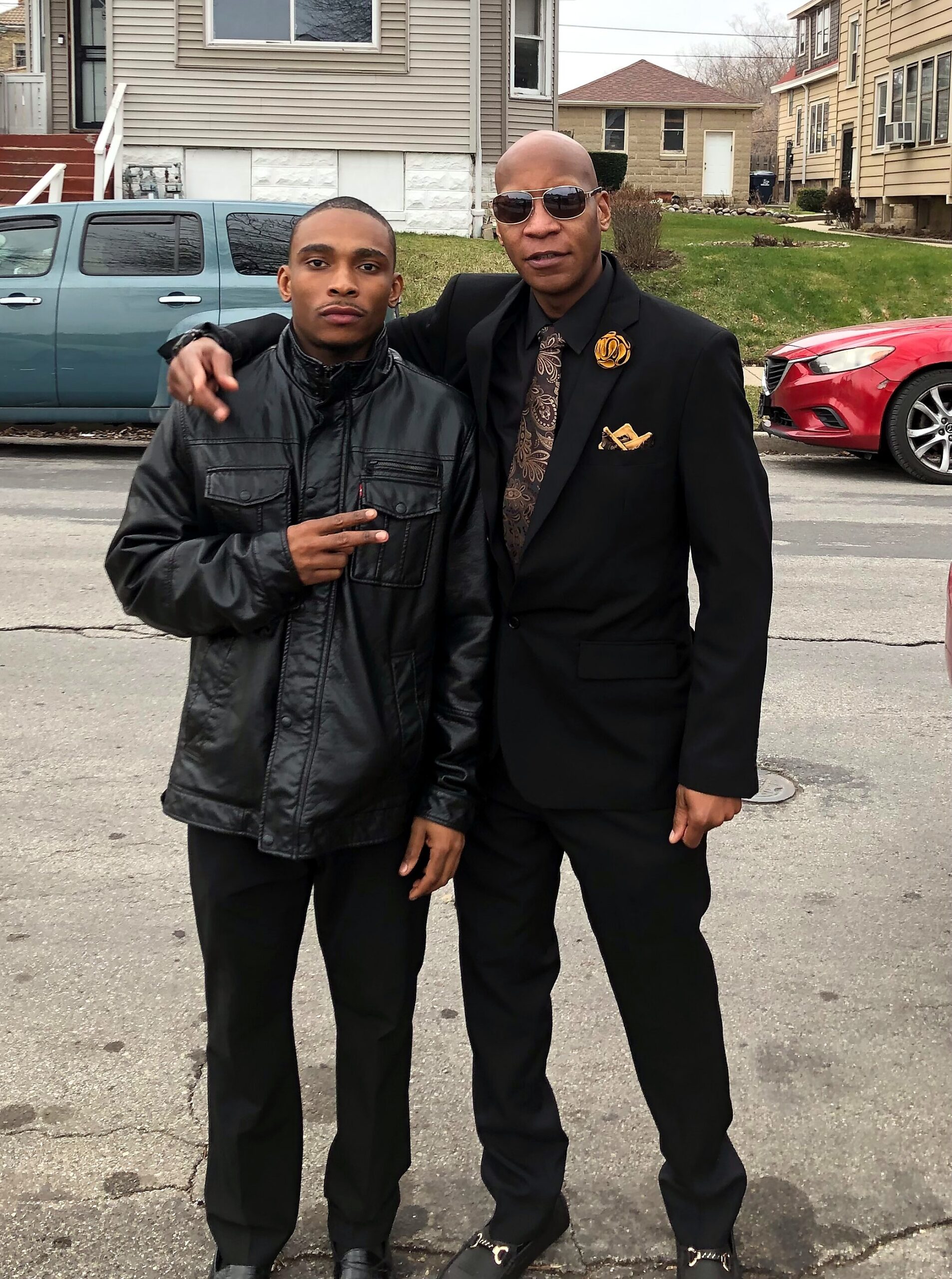 Two men pose for a photo on a residential street; left is in a black leather jacket making a peace sign, right wears a black suit and sunglasses with a gold lapel pin.