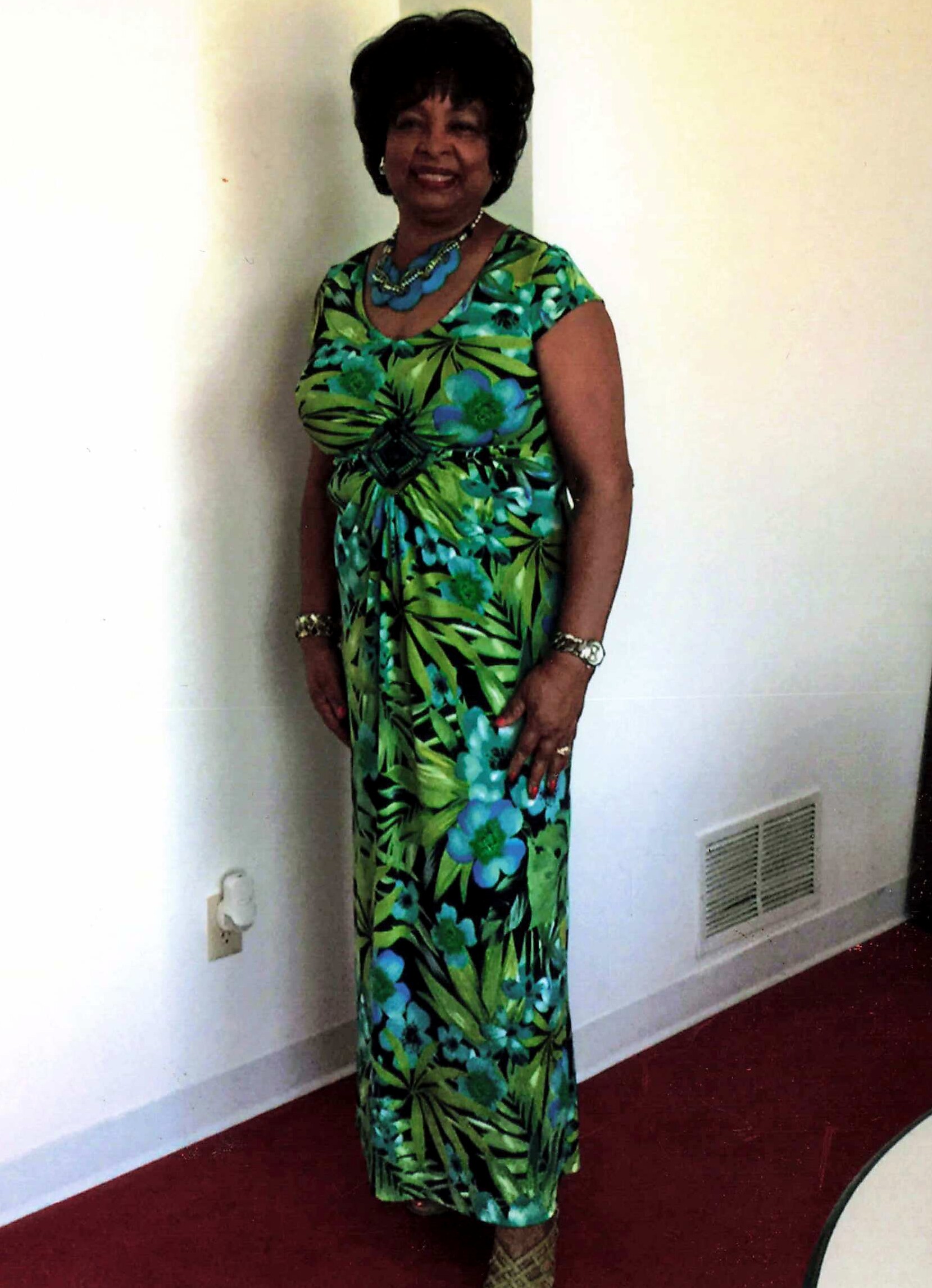 Woman in a green tropical-print dress standing indoors, smiling, wearing a blue statement necklace and bracelets.
