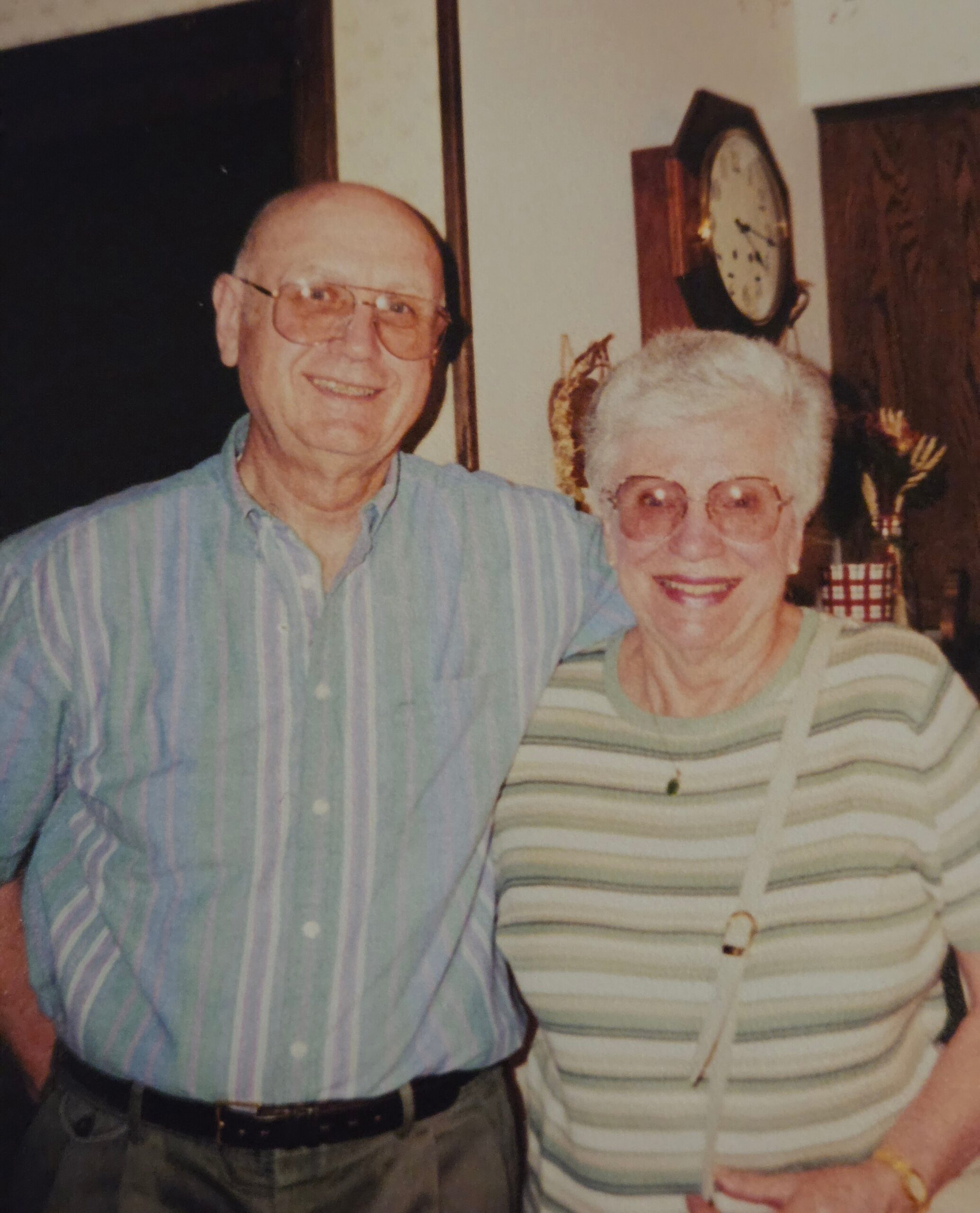 Smiling elderly man and woman posing indoors, man in a blue striped shirt and glasses, woman in a striped sweater.