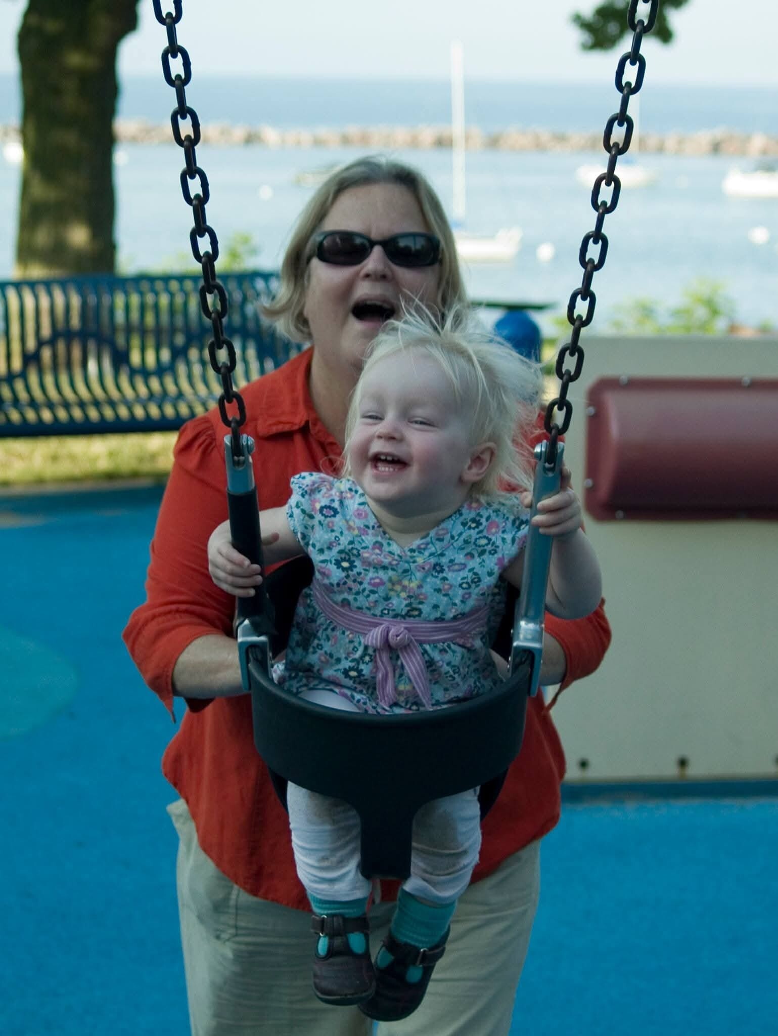 Woman in sunglasses pushes a laughing toddler on a swing at a seaside playground, blue ground and water in the background while boats float nearby.
