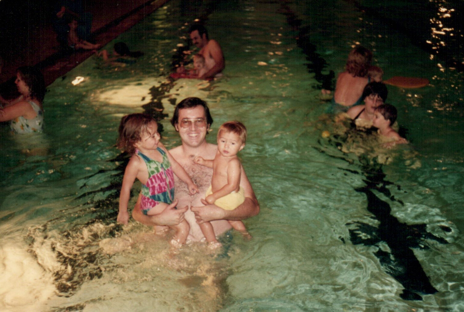 Man in a swimming pool holding two young children, one on each arm, with other swimmers in the background at night.
