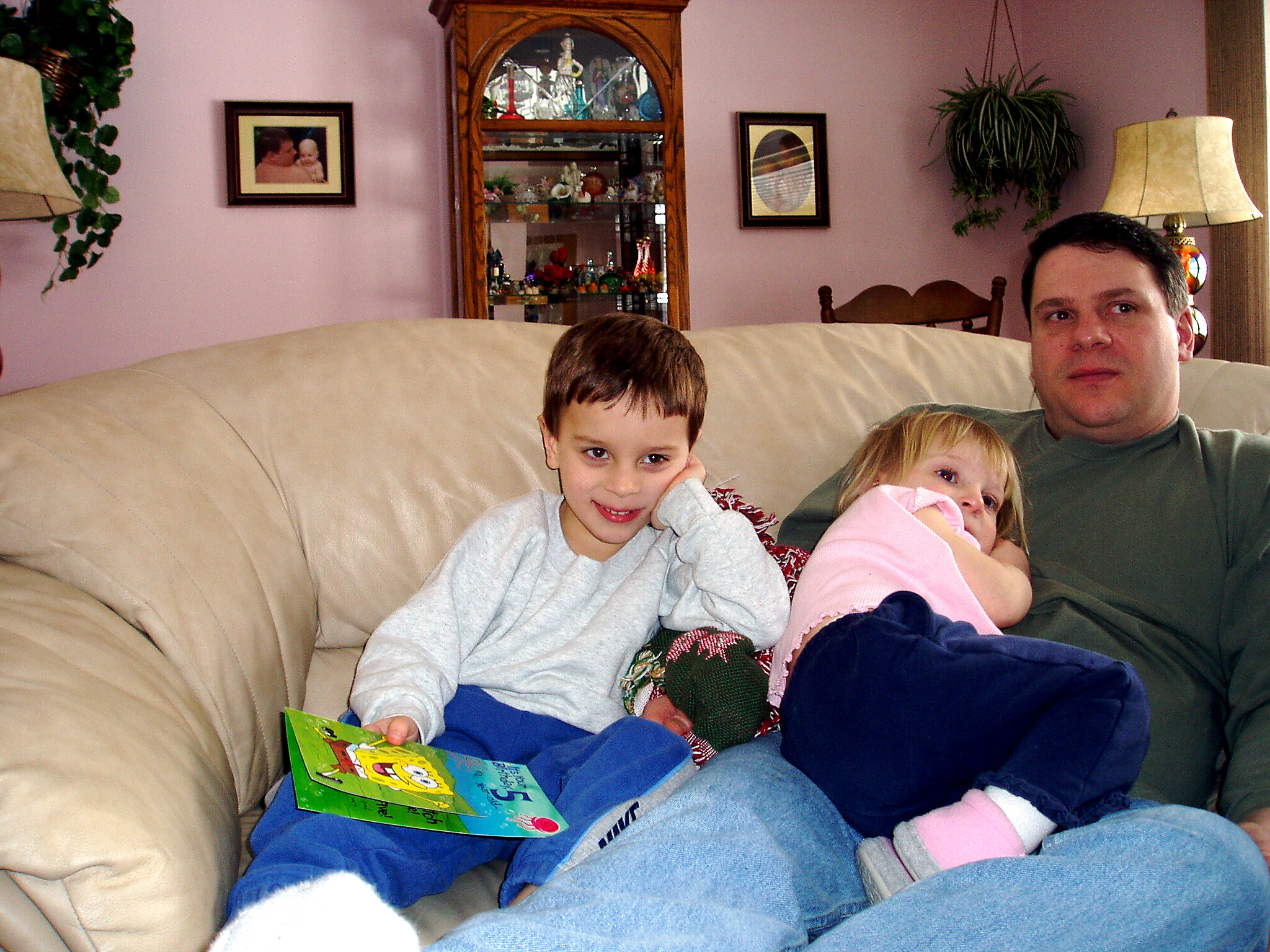 Father and two kids on a beige sofa in a pink-walled living room, with a colorful book in the boy's hand.