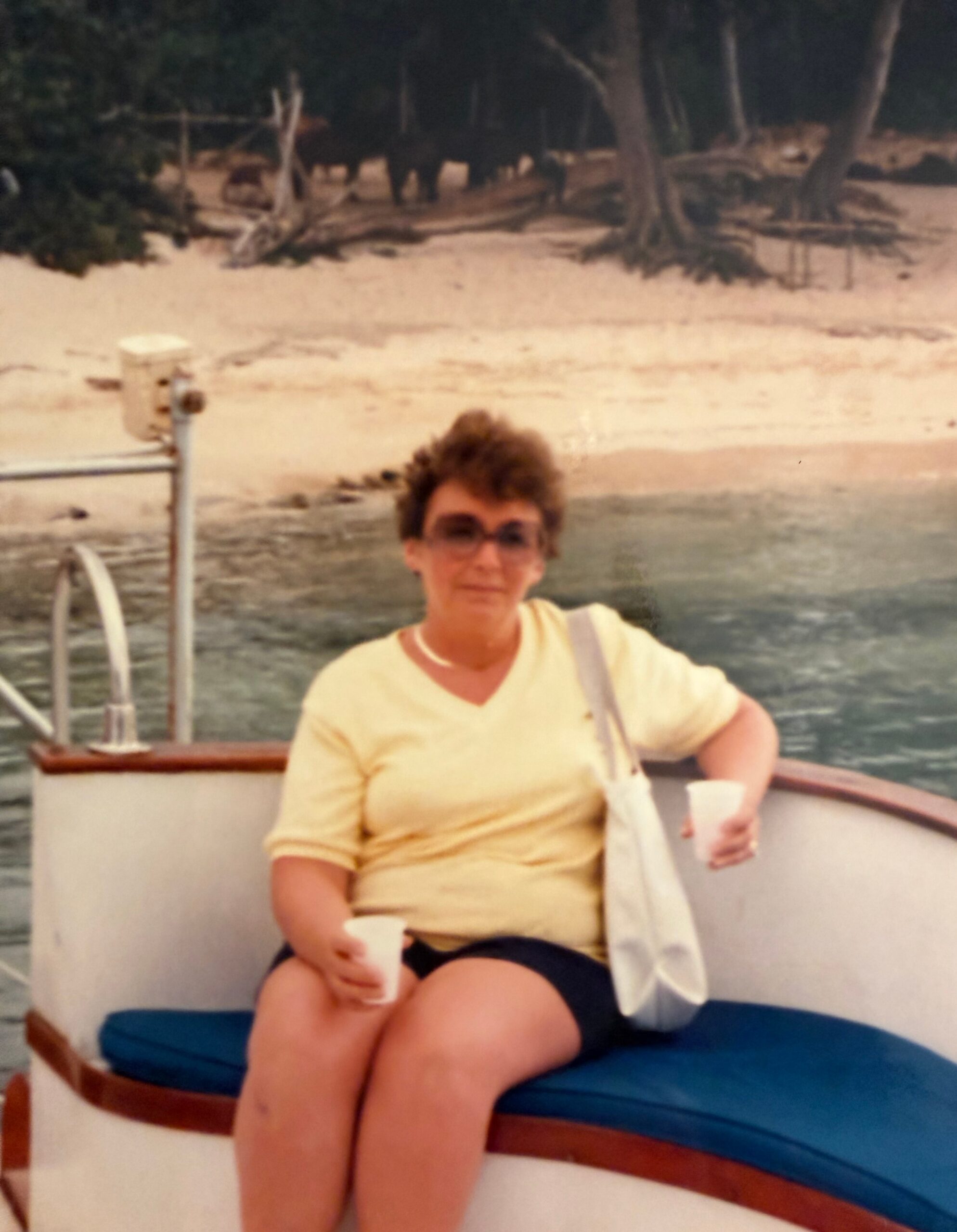 Person sitting on a boat by the water, wearing a light yellow shirt and holding a cup with a bag over the shoulder, with trees and shore in the background