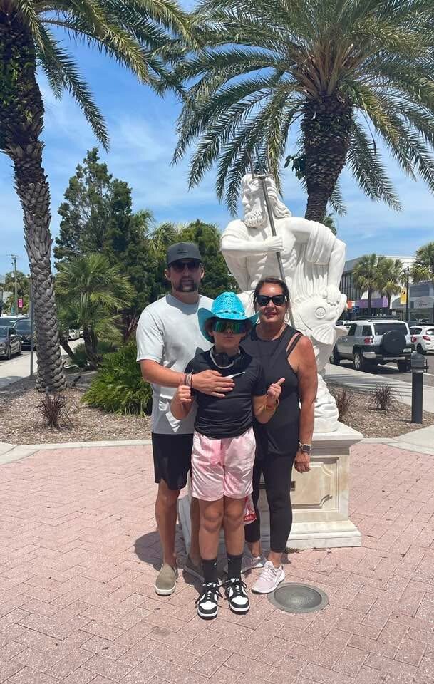 Family of three posing in front of a white statue outdoors, with palm trees and parked cars in a sunny plaza behind them.