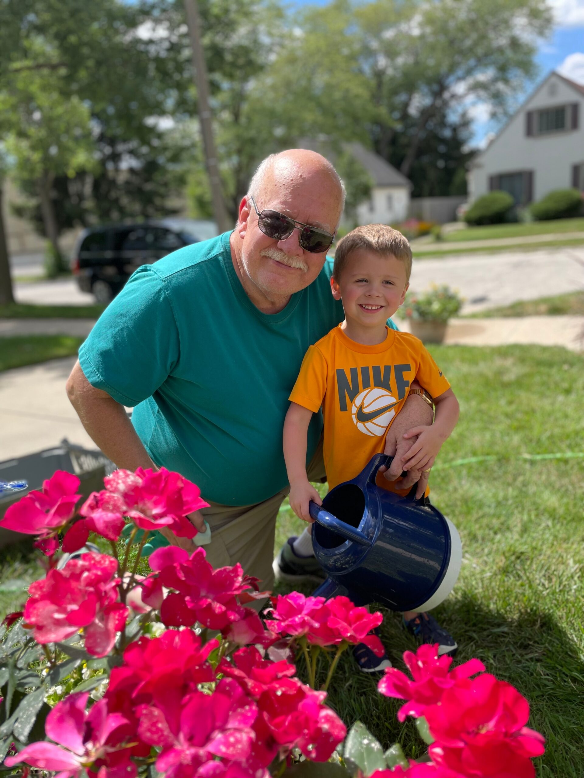 Grandfather and grandson smiling while watering pink flowers in a sunny front yard.