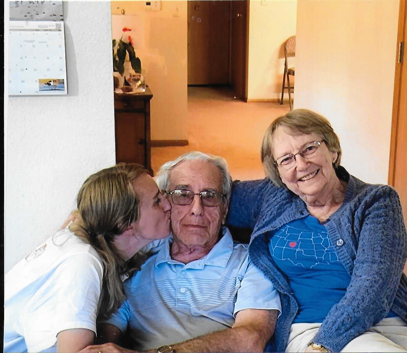 An elderly man with glasses sits between two smiling women; the left woman kisses his cheek, the right woman smiles at the camera in a home setting.