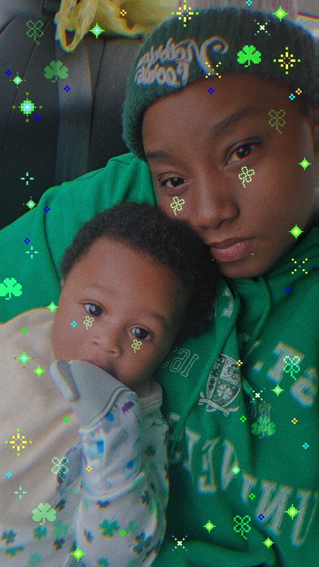 Teenager in a green hoodie cradling a baby in a car, both looking at the camera, with a colorful glitter filter overlaid.