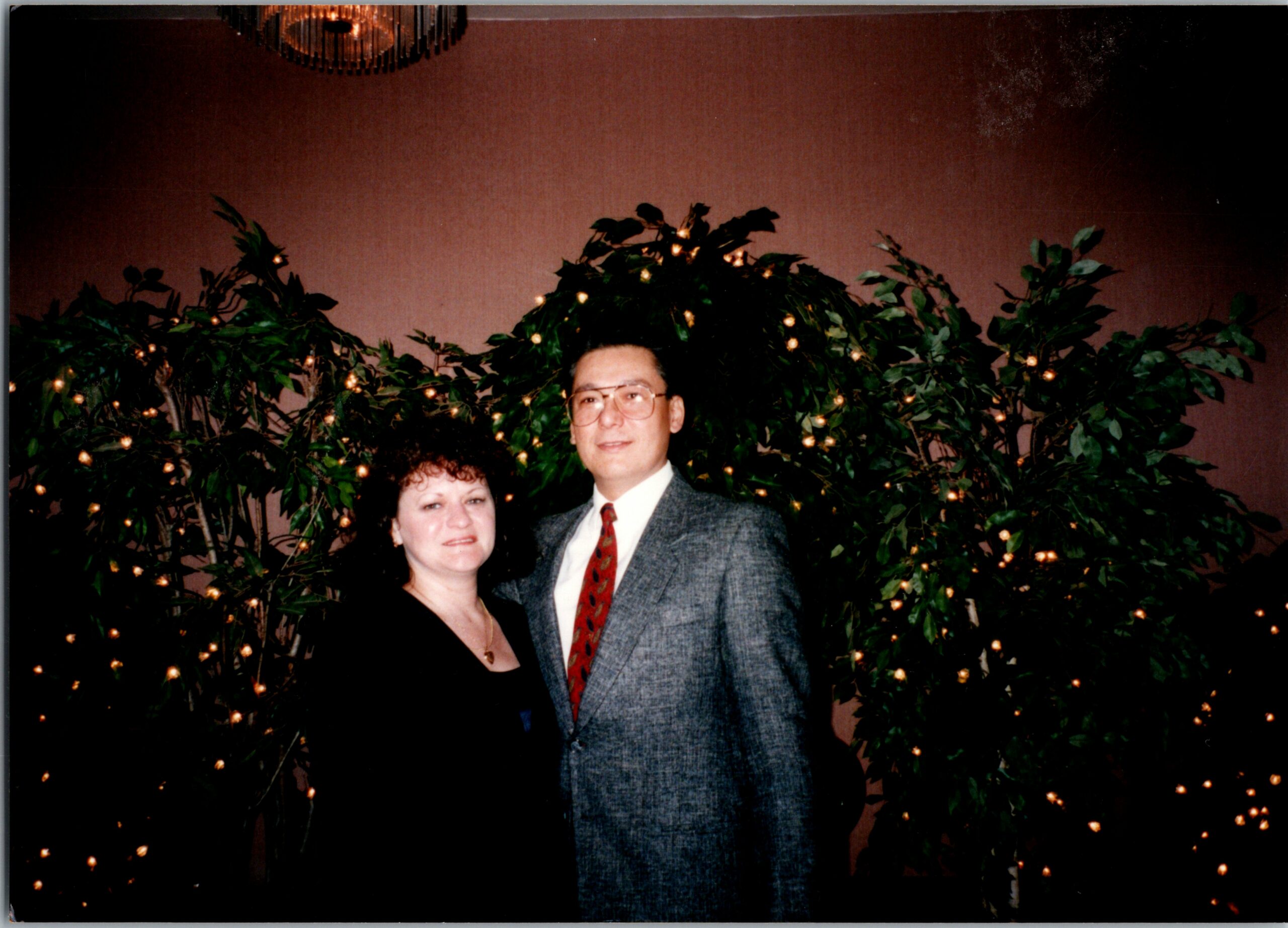 Smiling couple in formal attire posing in front of leafy backdrop with twinkling lights