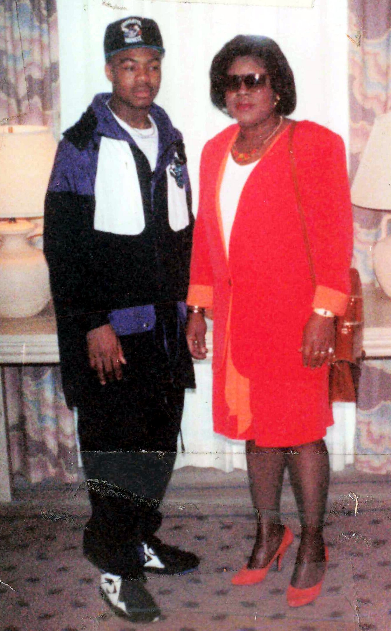 Two people posing indoors: a man in a black/purple jacket and cap beside a woman in a bright red suit with sunglasses and jewelry.