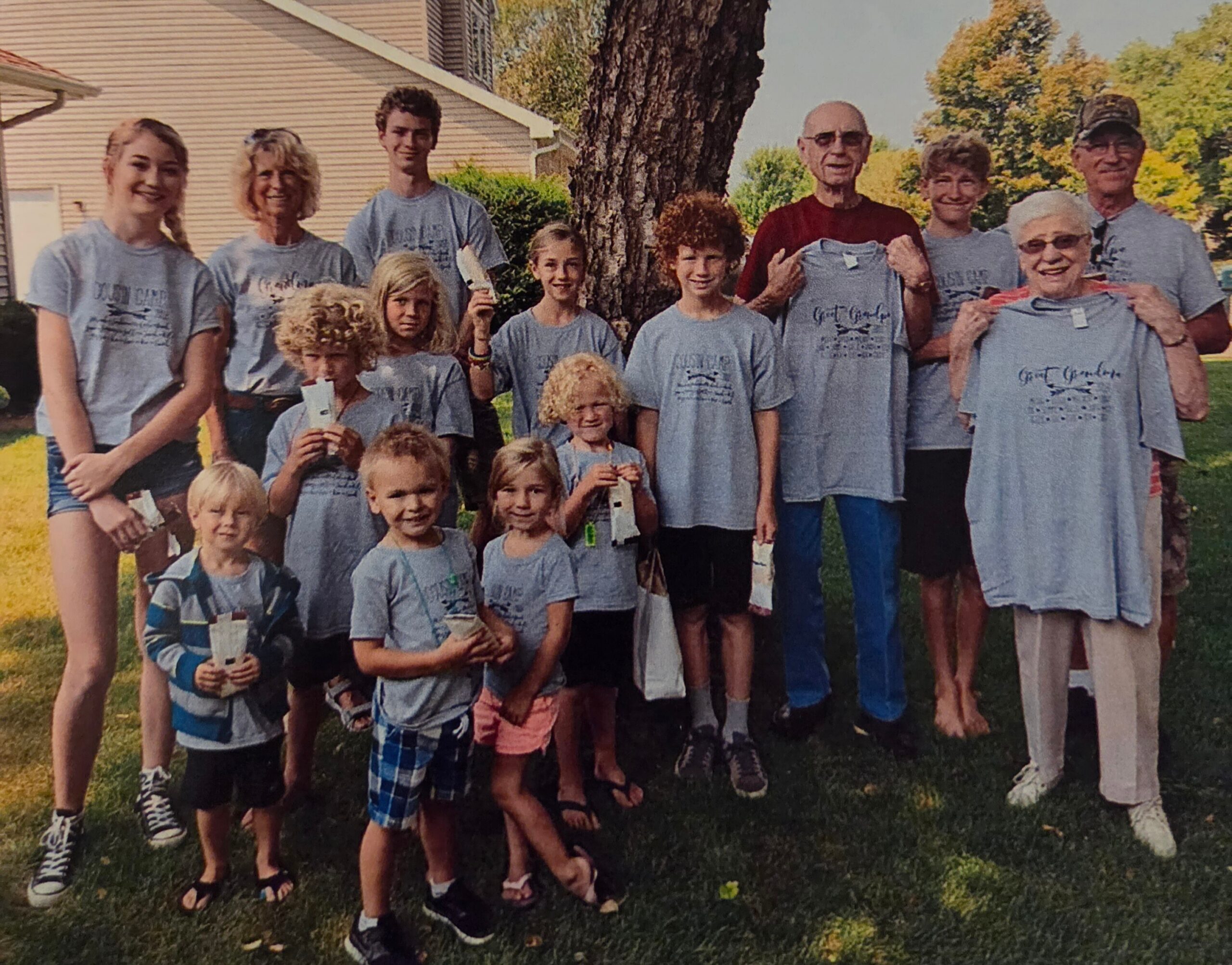 Group of children and adults wearing matching gray T-shirts pose together on a lawn with a tree and house in the background, smiling for a photo.
