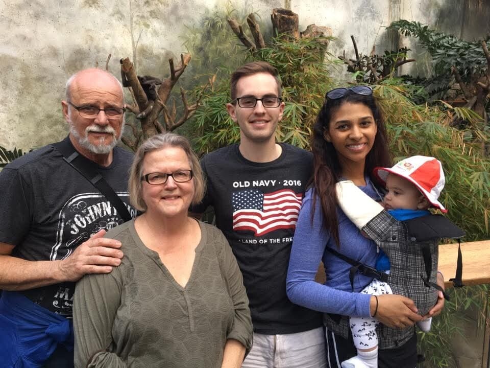 Group of five posing outdoors: an older man and woman with two younger adults and a baby in a carrier.