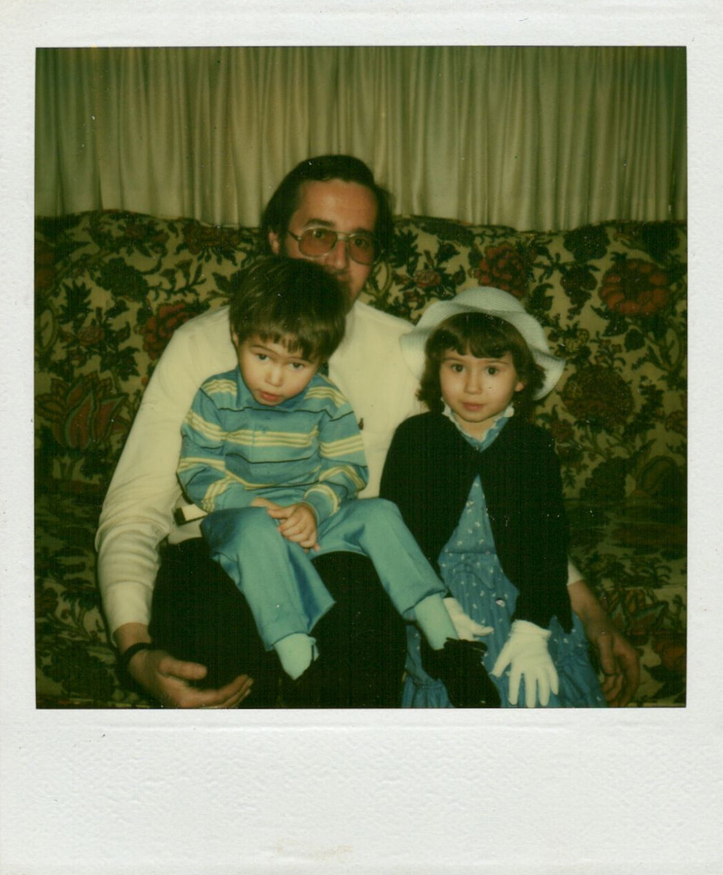 Father with two children posing on a floral patterned couch; boy in blue striped shirt sits on his lap, girl in blue dress with a white hat beside him.