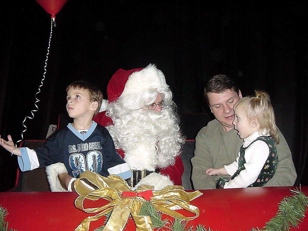 Santa Claus sits with a boy and a girl on a stage, a man holds the girl nearby, with a gold ribbon bow in the foreground.