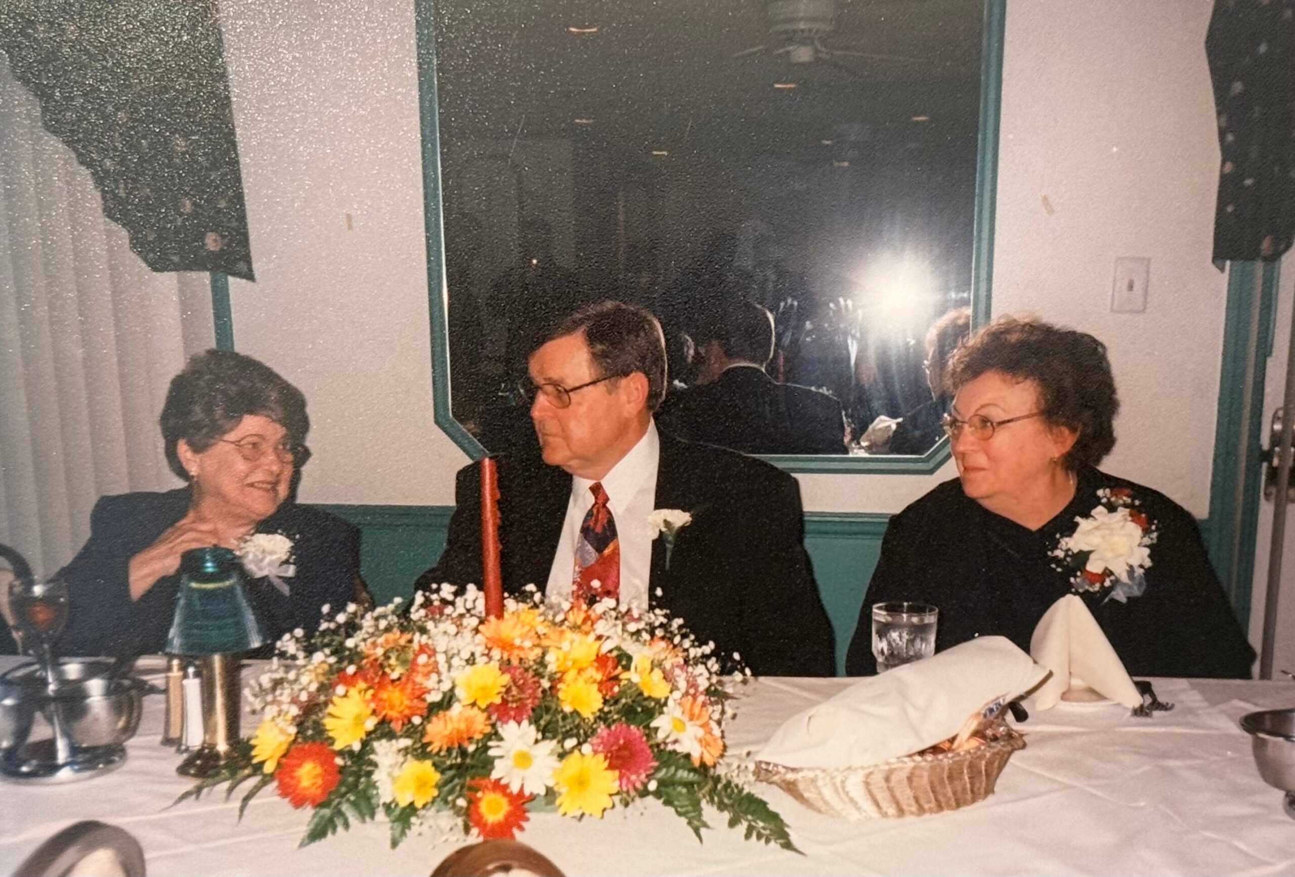Three older adults in formal attire sit at a wedding reception table, with a colorful flower centerpiece in front of them.
