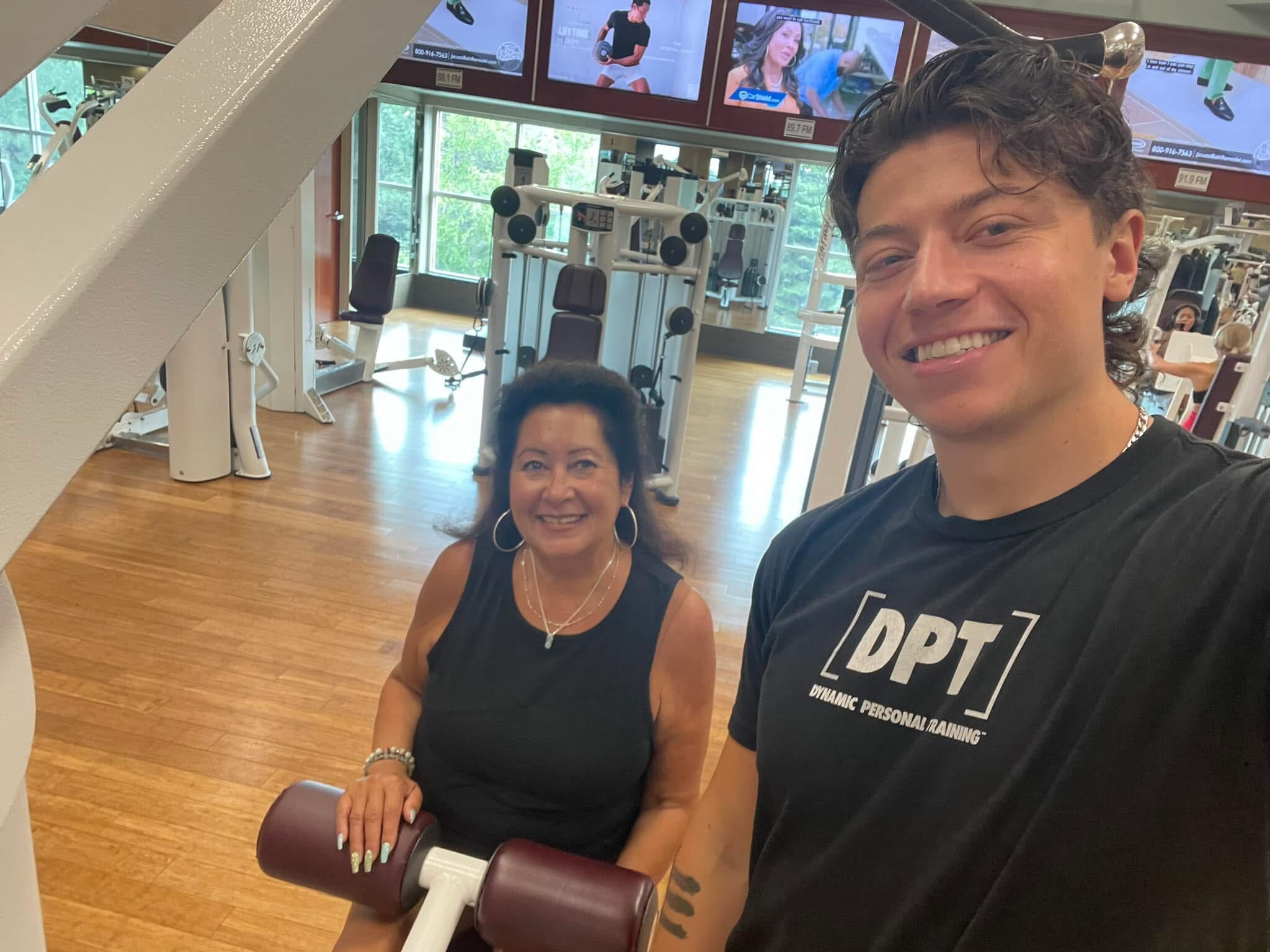Smiling man in a black DPT shirt taking a selfie with a woman in a black top at the gym, standing next to a core roller and weight machines in the background.
