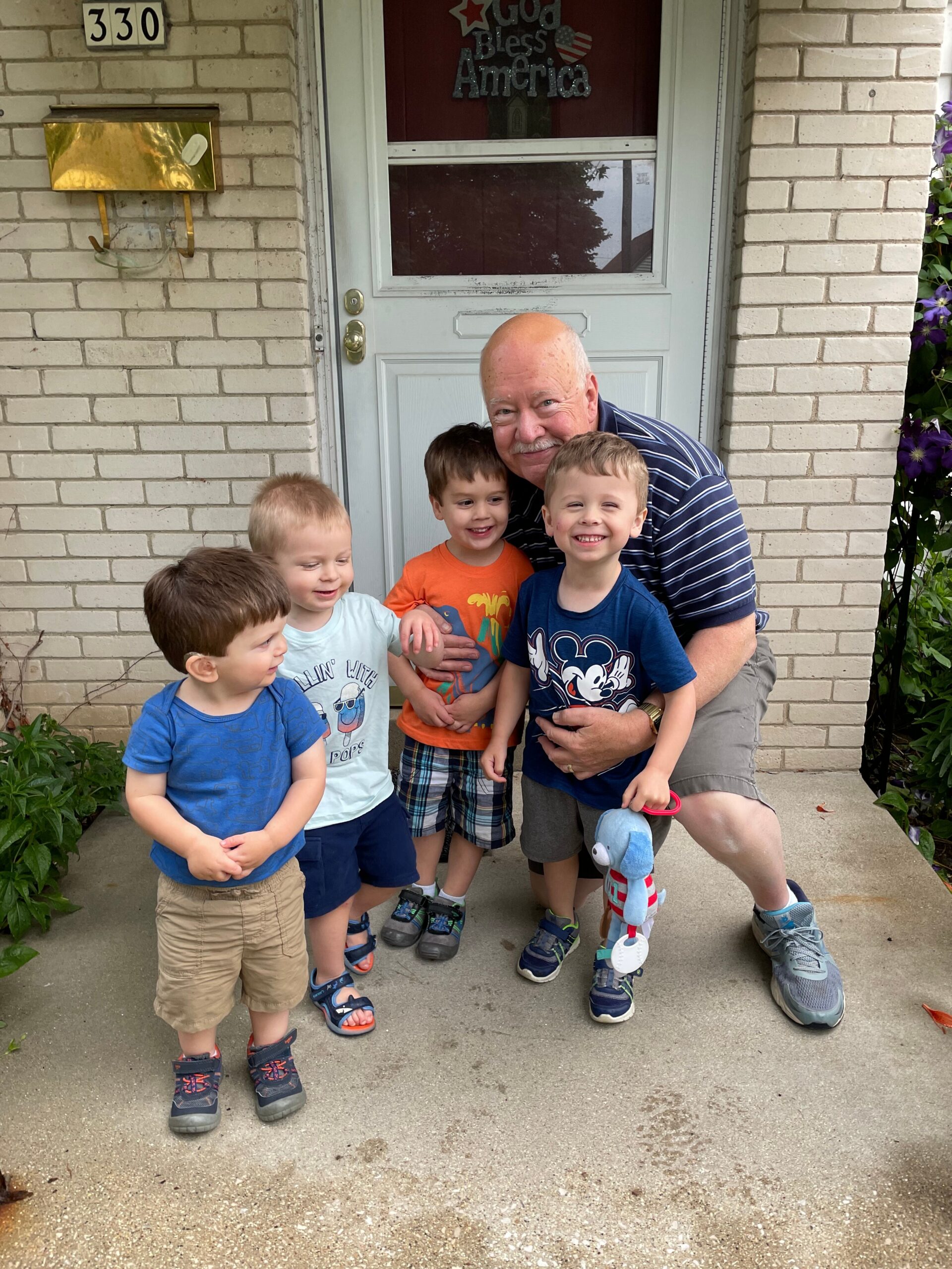 Grandfather with four smiling kids posing together on a porch in front of a white brick house door.