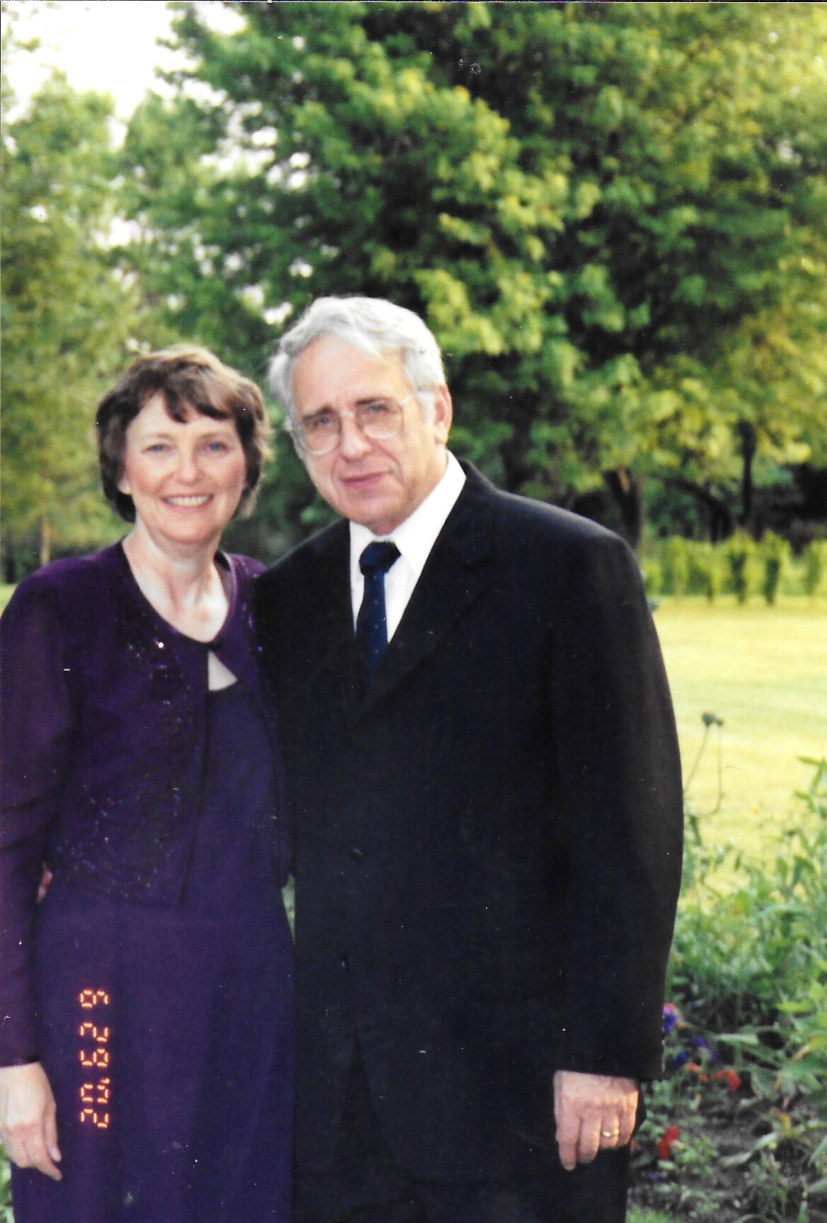 Older couple posing together outdoors in a garden, smiling; woman wears a purple dress and jacket, man in a dark suit and tie, with trees and flowers in the background.