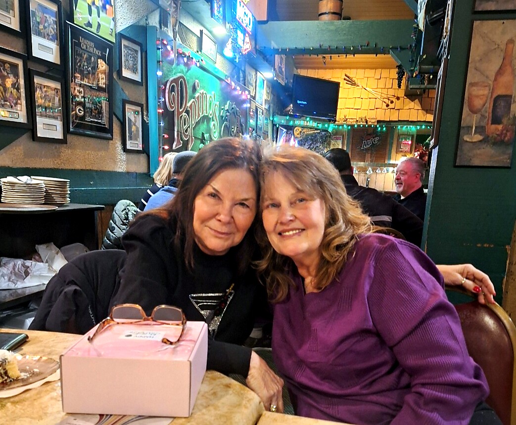 Two women smiling at a restaurant table, pink gift box with sunglasses in the foreground and colorful neon signs in the background on the wall.