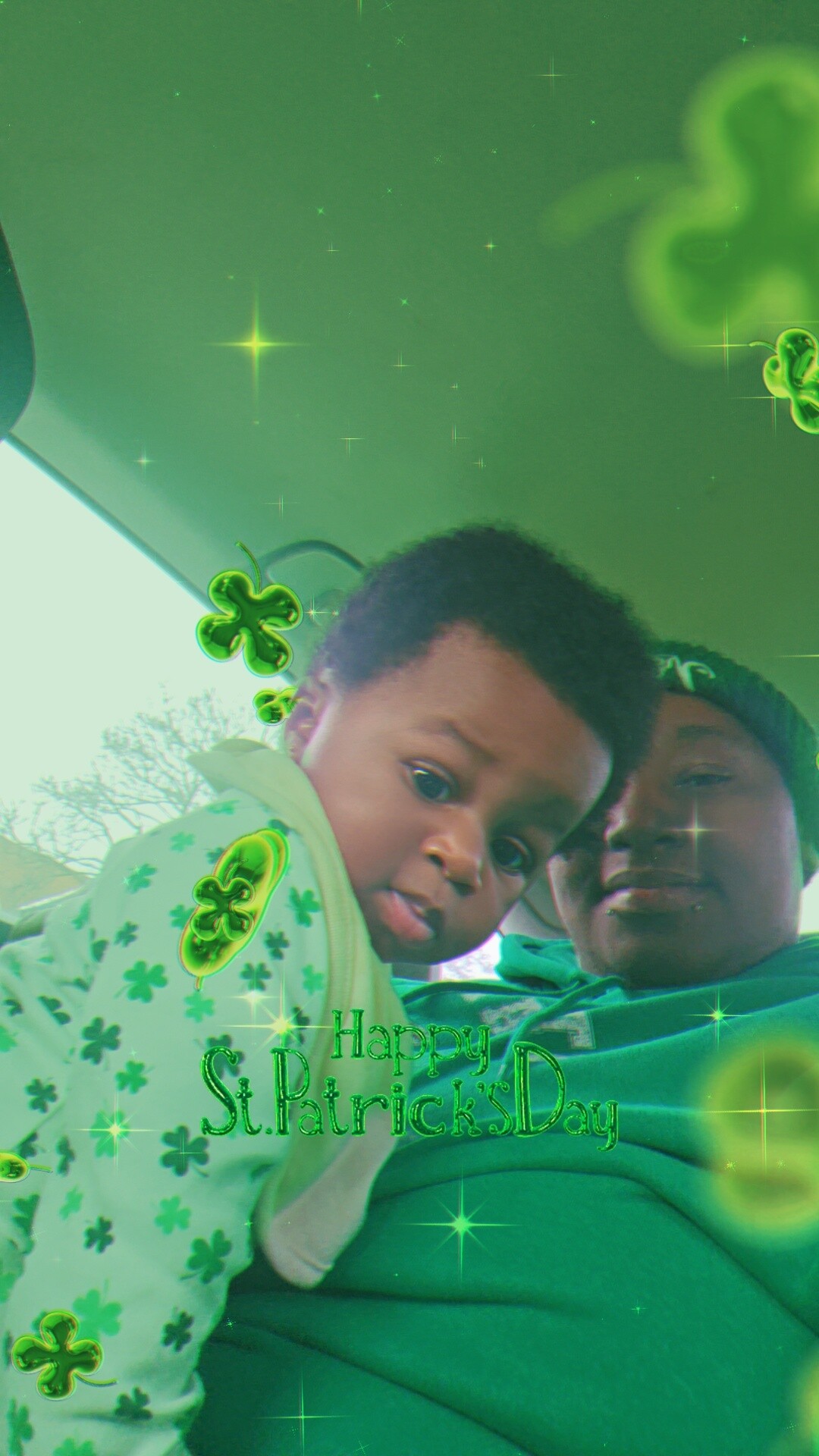 Two children in a car wearing green clothing, with St. Patrick's Day shamrock decorations and a green glow overlay