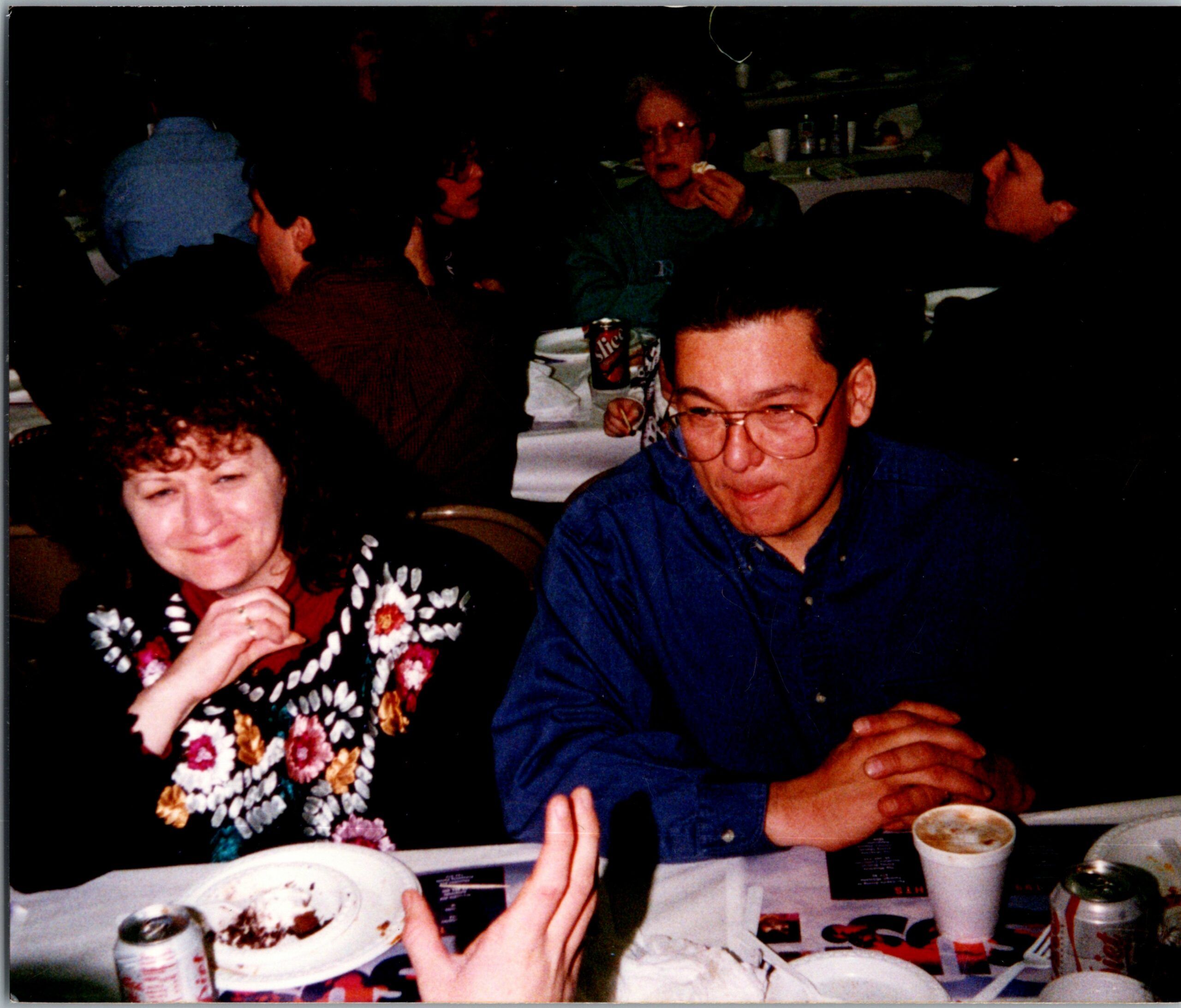 People dining at a crowded table, talking and eating; foreground shows a woman in a floral cardigan and a man in a blue shirt.