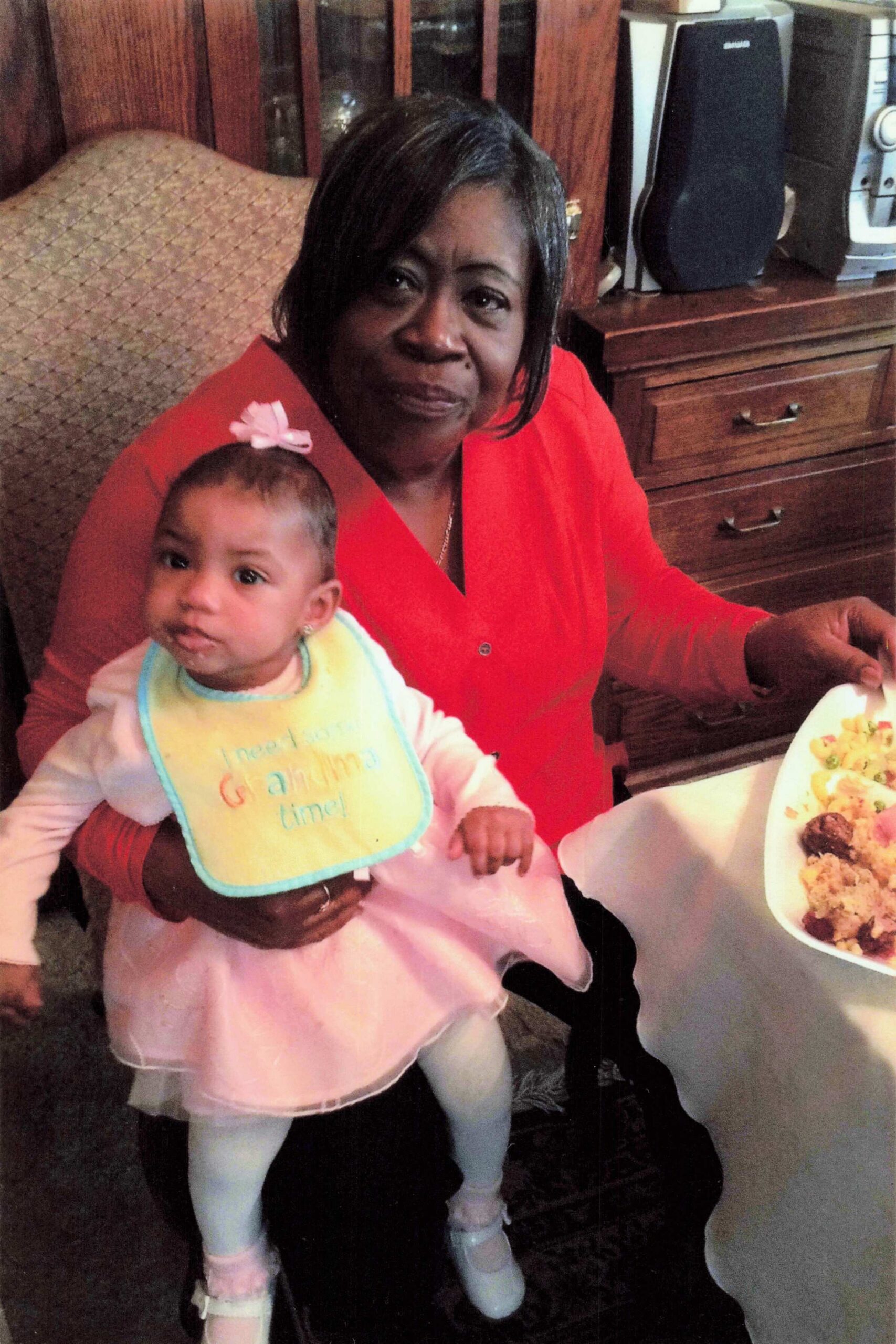 Smiling woman in a red shirt holding a baby girl in a pink dress with a bow, seated at a dining table with food nearby.