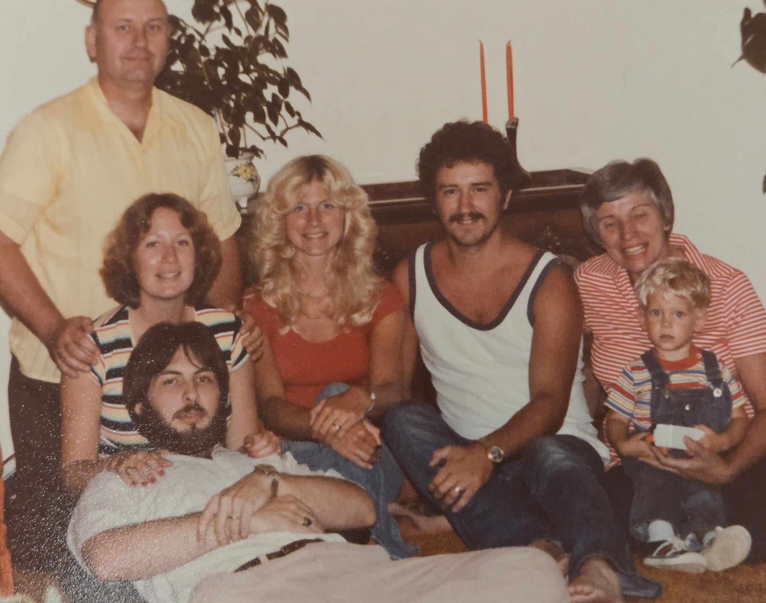 Group of adults and a young child posing together in a living room, smiling for a casual family photo in retro clothing and setting.