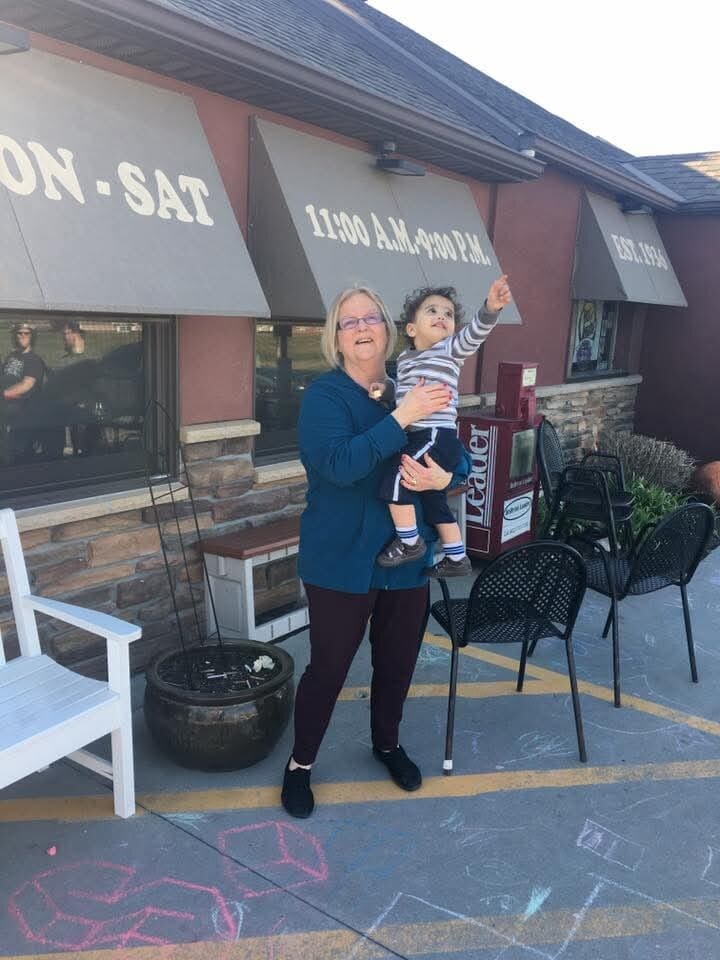 Woman in a blue jacket holds a smiling toddler in striped clothes outside a cafe with hour signs on the awnings.