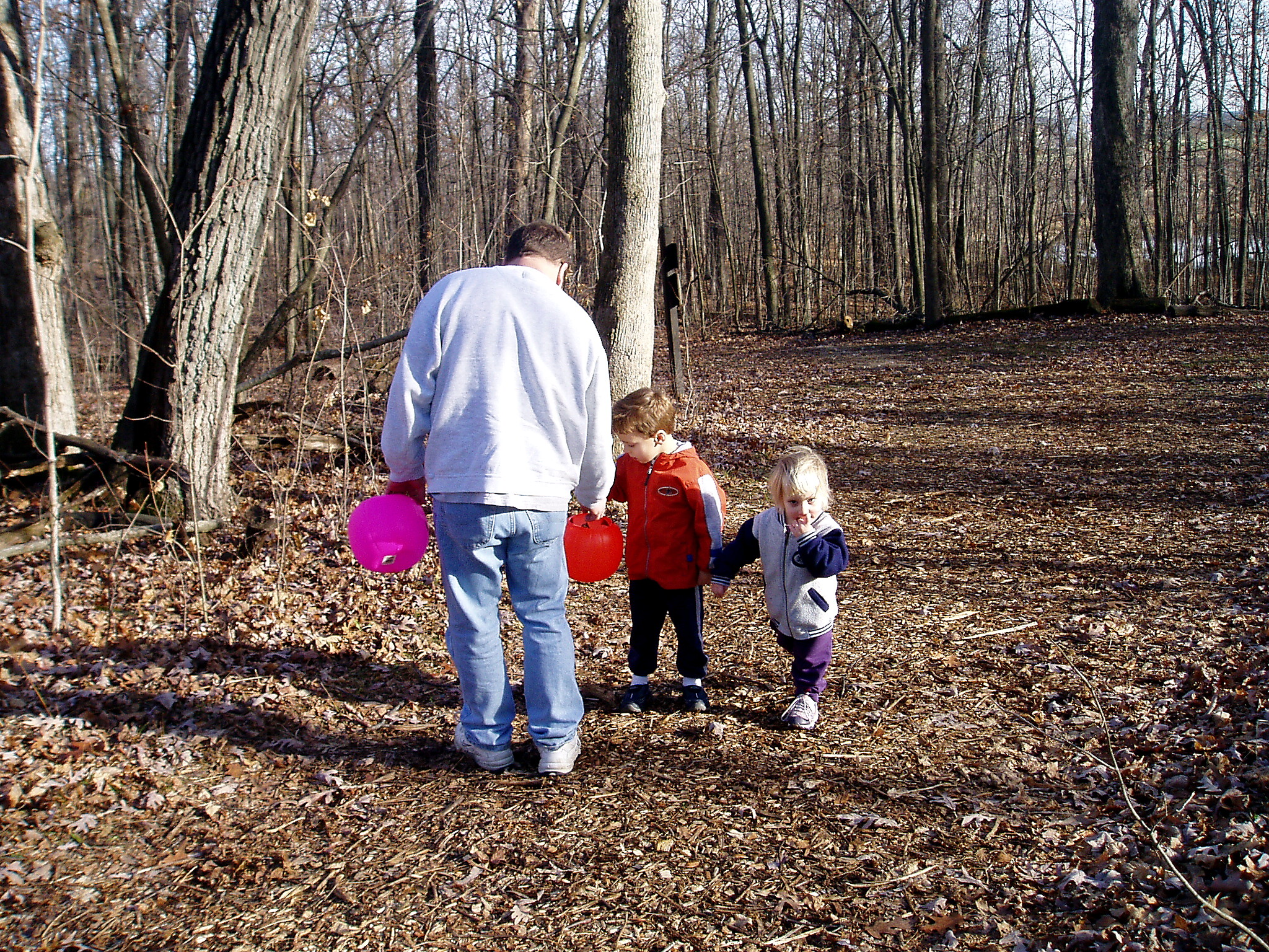 Adult man in a gray hoodie stands with two young children in a leaf-covered forest, each child holding a colorful balloon (pink and orange). Two adults/children pose near bare trees on a sunny day.