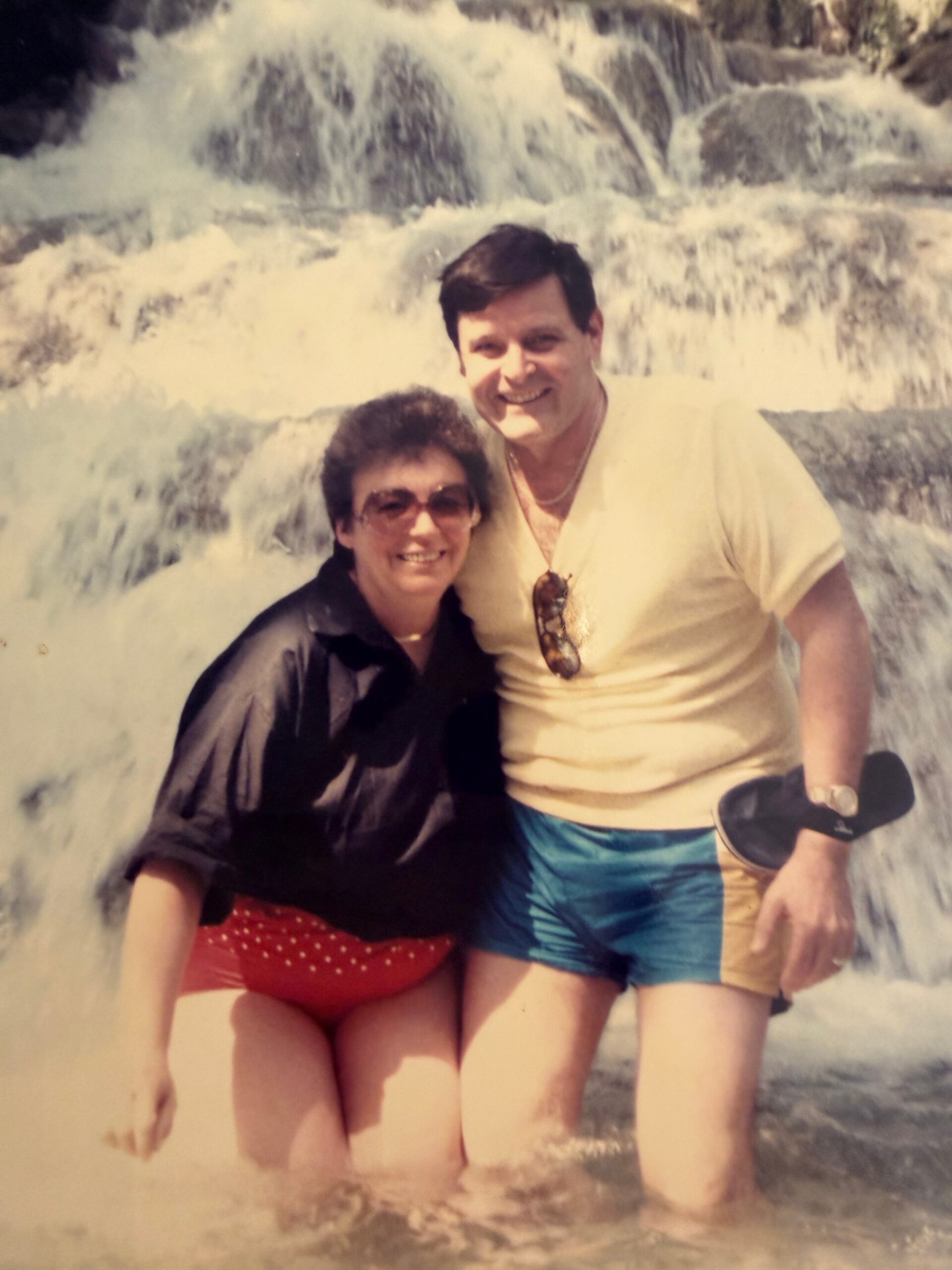 Couple posing in front of a cascading waterfall, smiling, wearing swimsuits; woman in red polka-dot bottoms, man in yellow shirt and blue shorts.