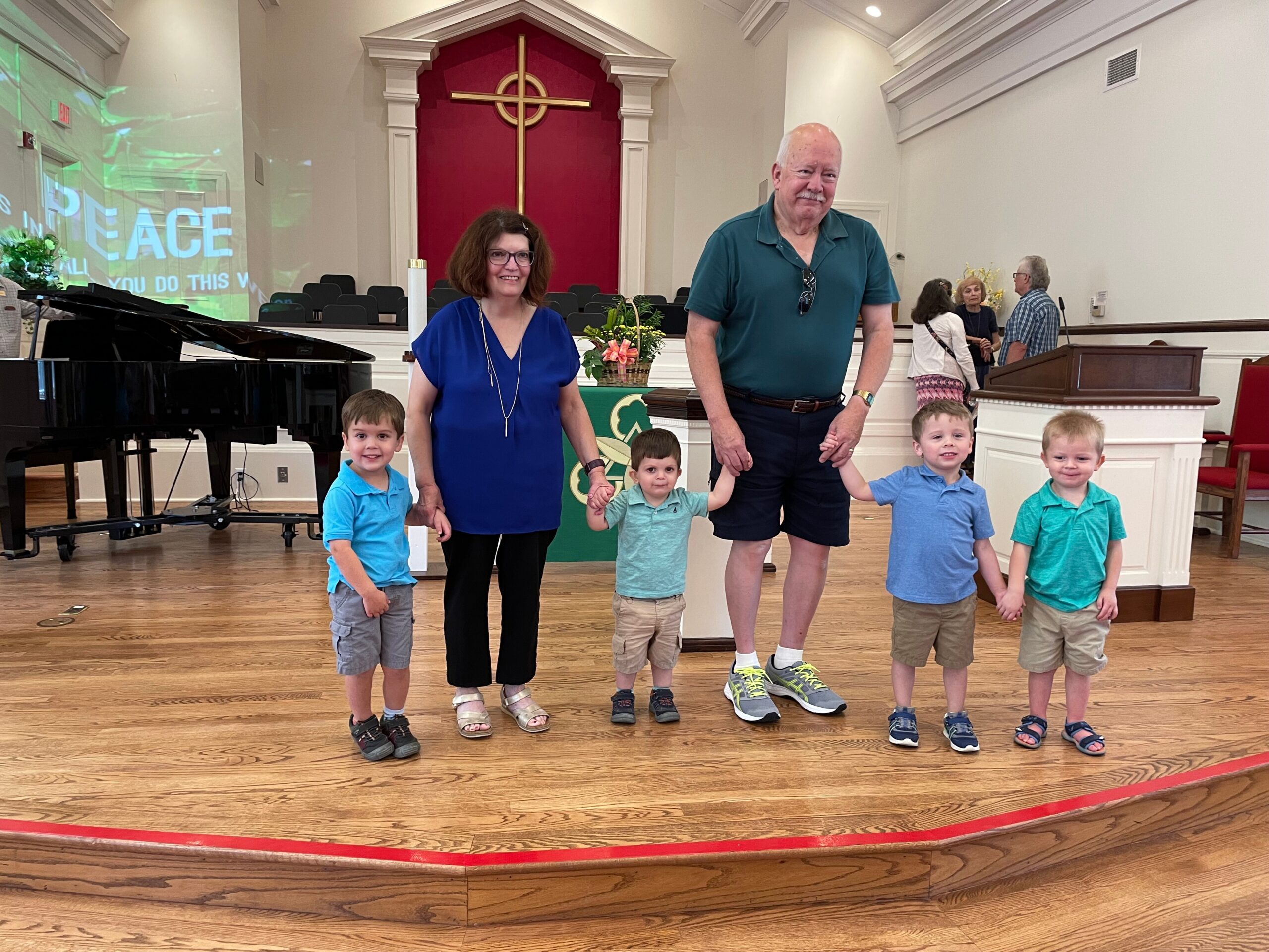 Family and adults posing on a church stage in front of a red altar with a gold cross; several young children stand at the front, holding hands with the adults behind them.