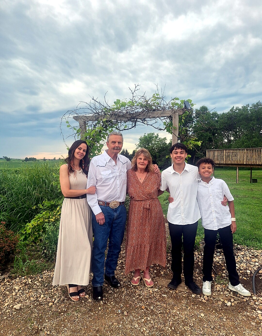 Five people posing together outdoors under a vine-covered wooden arch in a garden.