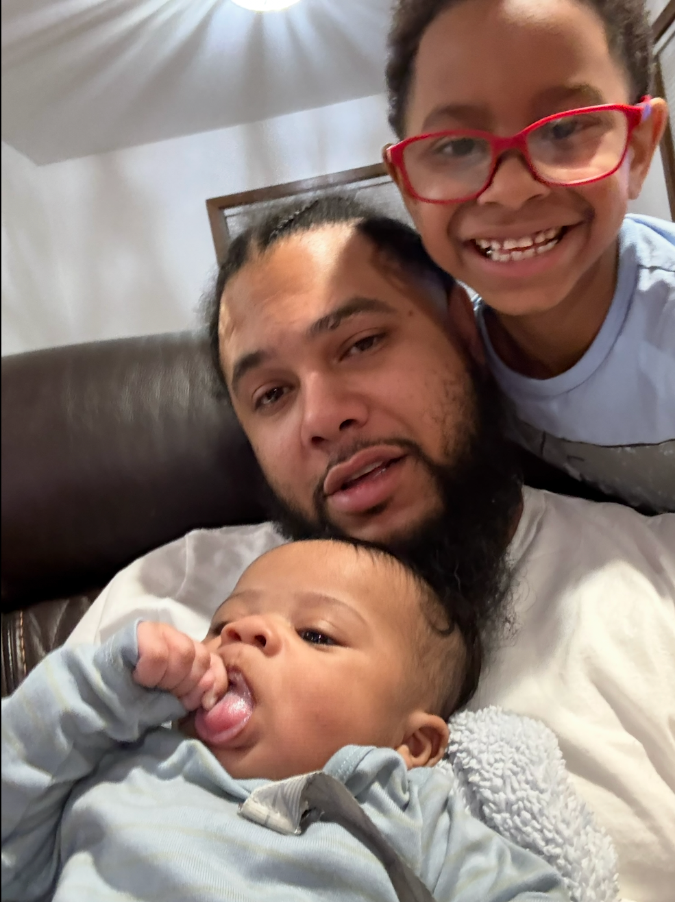 Father with a beard lying on a sofa, holding a baby while an older child with red glasses leans in smiling nearby