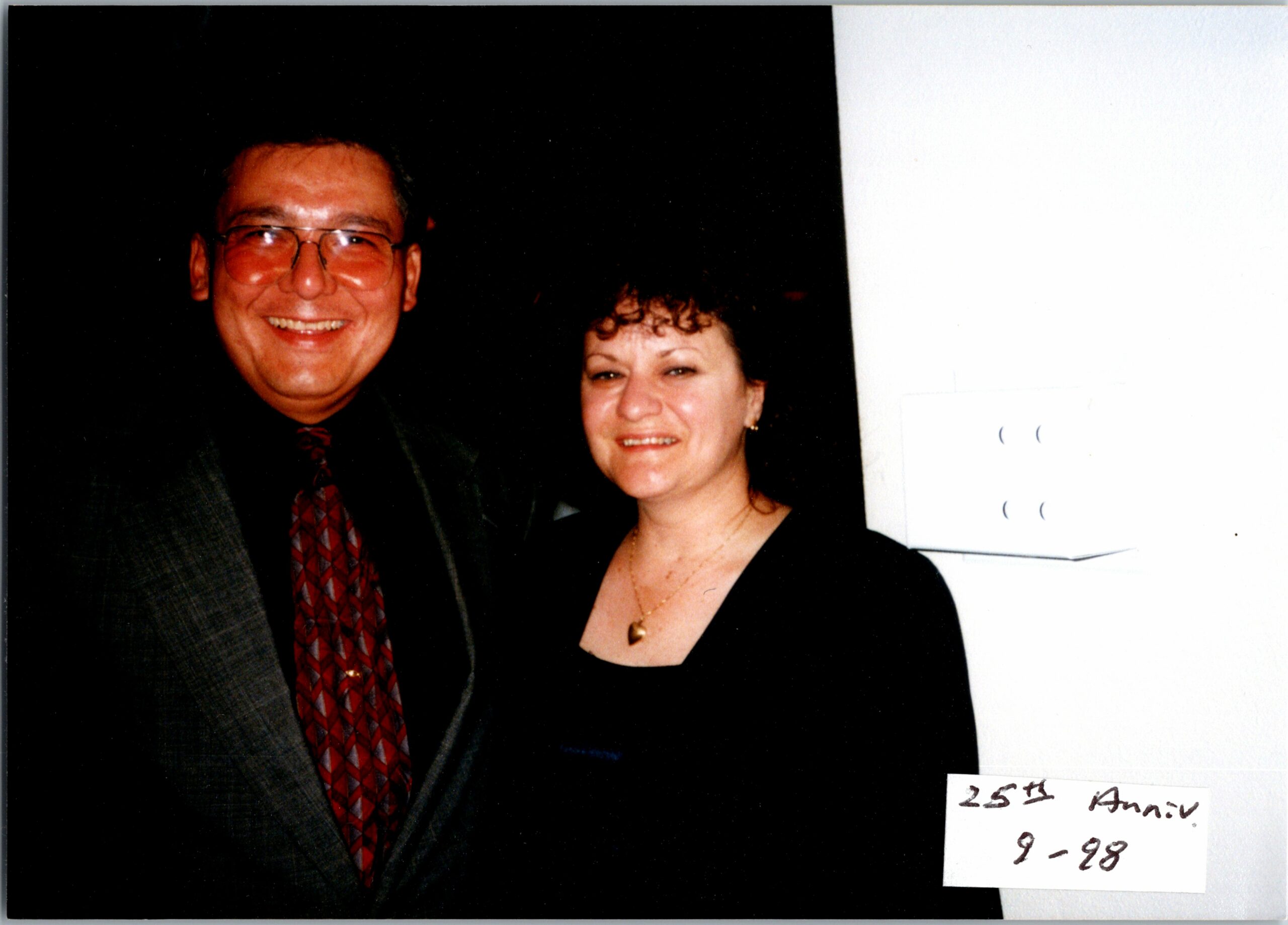 Smiling man in a dark suit and red patterned tie beside a woman in a black dress at a formal event.