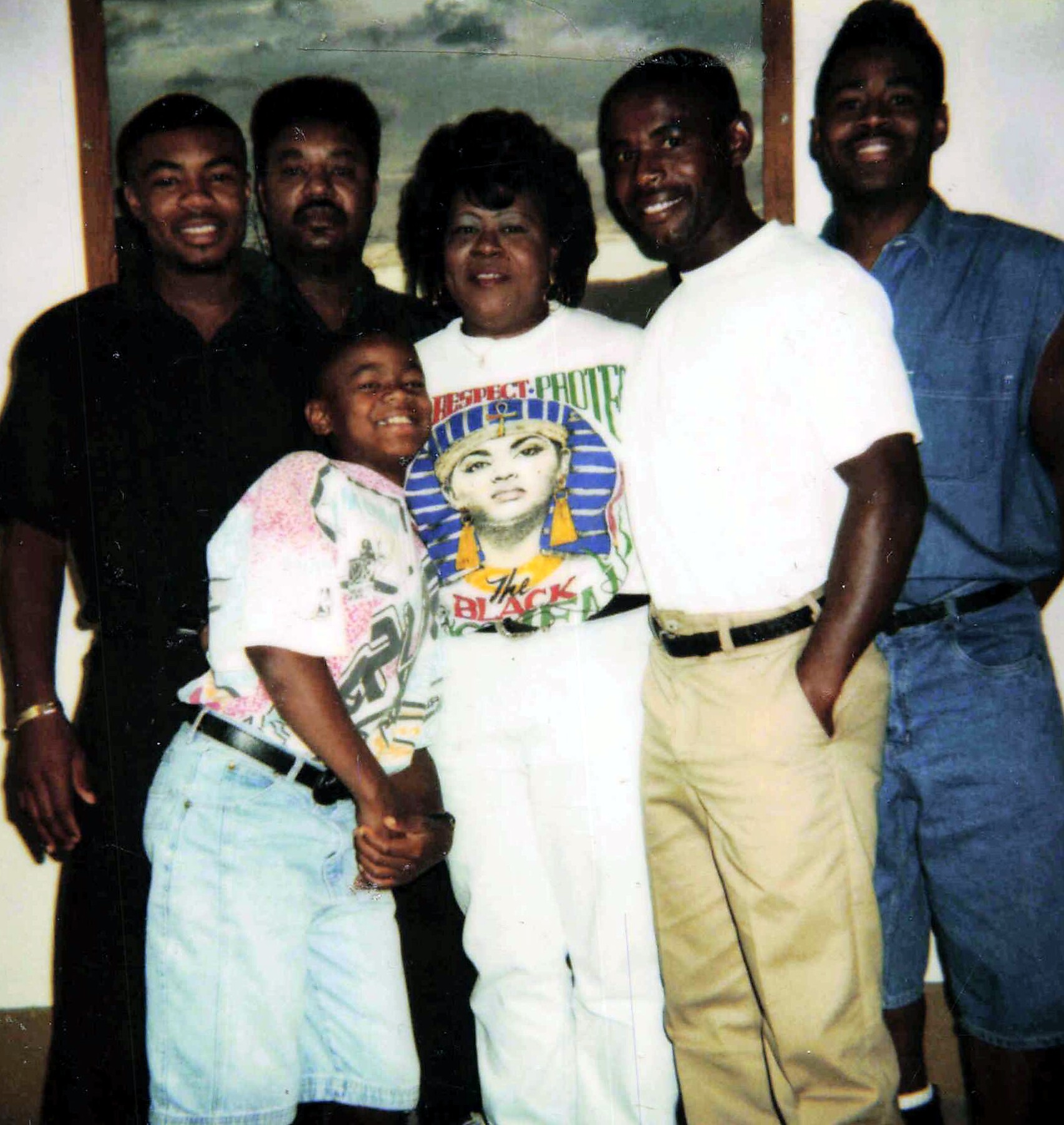 Group of six people posing indoors, smiling at the camera; a woman in a white shirt stands center with others around her, and a child sits in front wearing a light jacket.