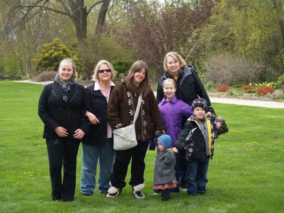 Seven people, adults and children, posing together on a park lawn with trees in the background.