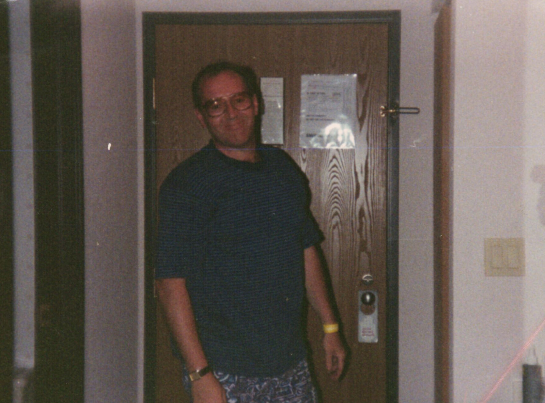 Man with glasses wearing a dark striped shirt and patterned shorts stands in a doorway inside a building.