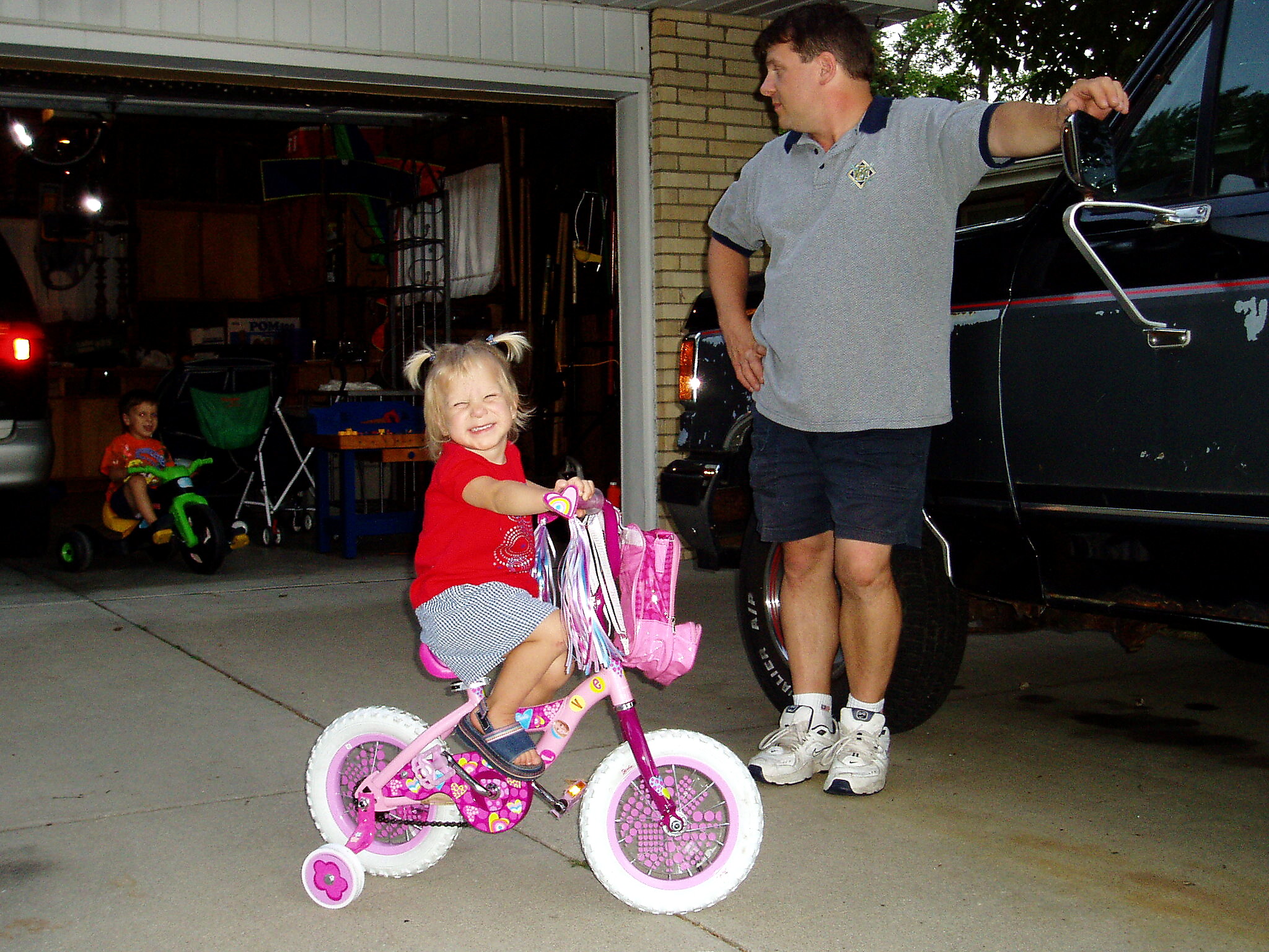 Young girl riding a pink bicycle with training wheels in a driveway, smiling as a man stands nearby by a parked van in the background.