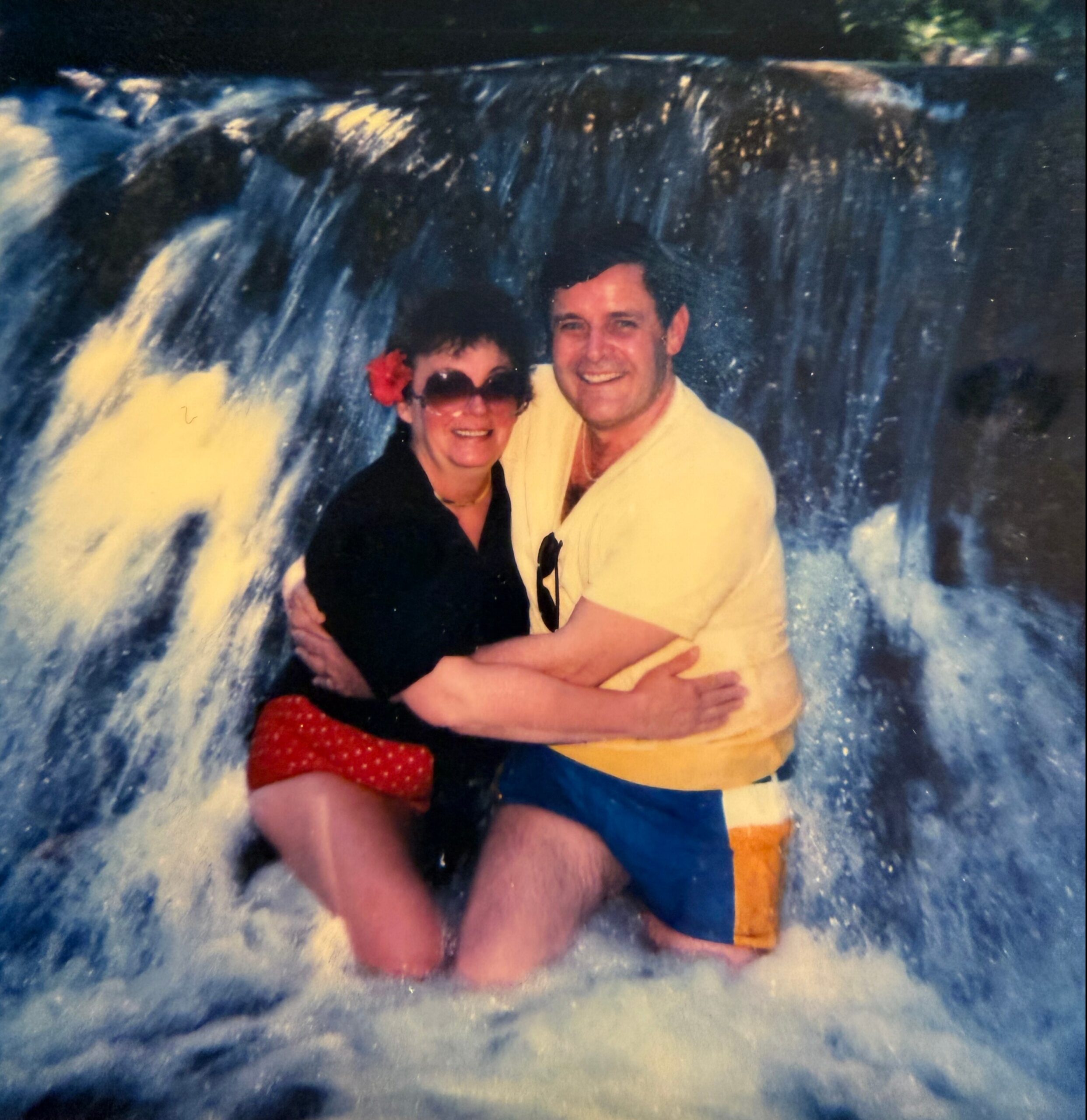 Couple posing together outdoors, smiling, with their arms around each other in front of a blue, water-splash background.