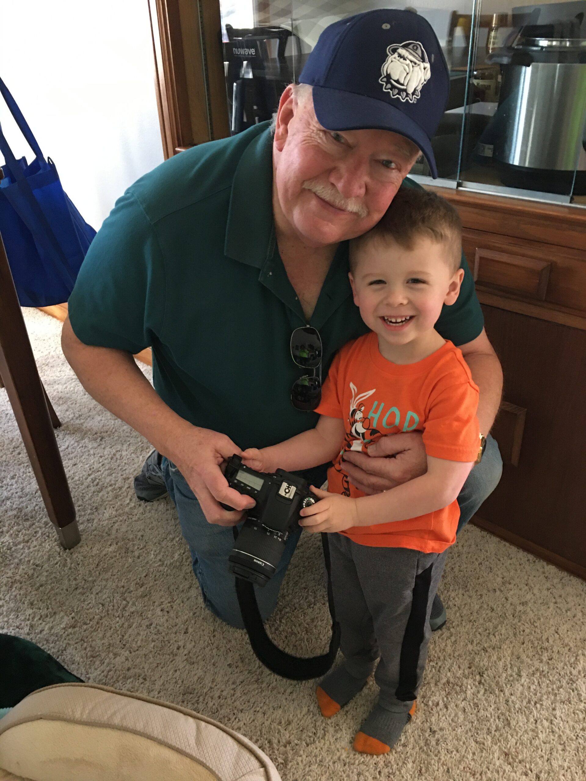 Older man kneeling beside a smiling child, holding a DSLR camera with a strap in a home setting.