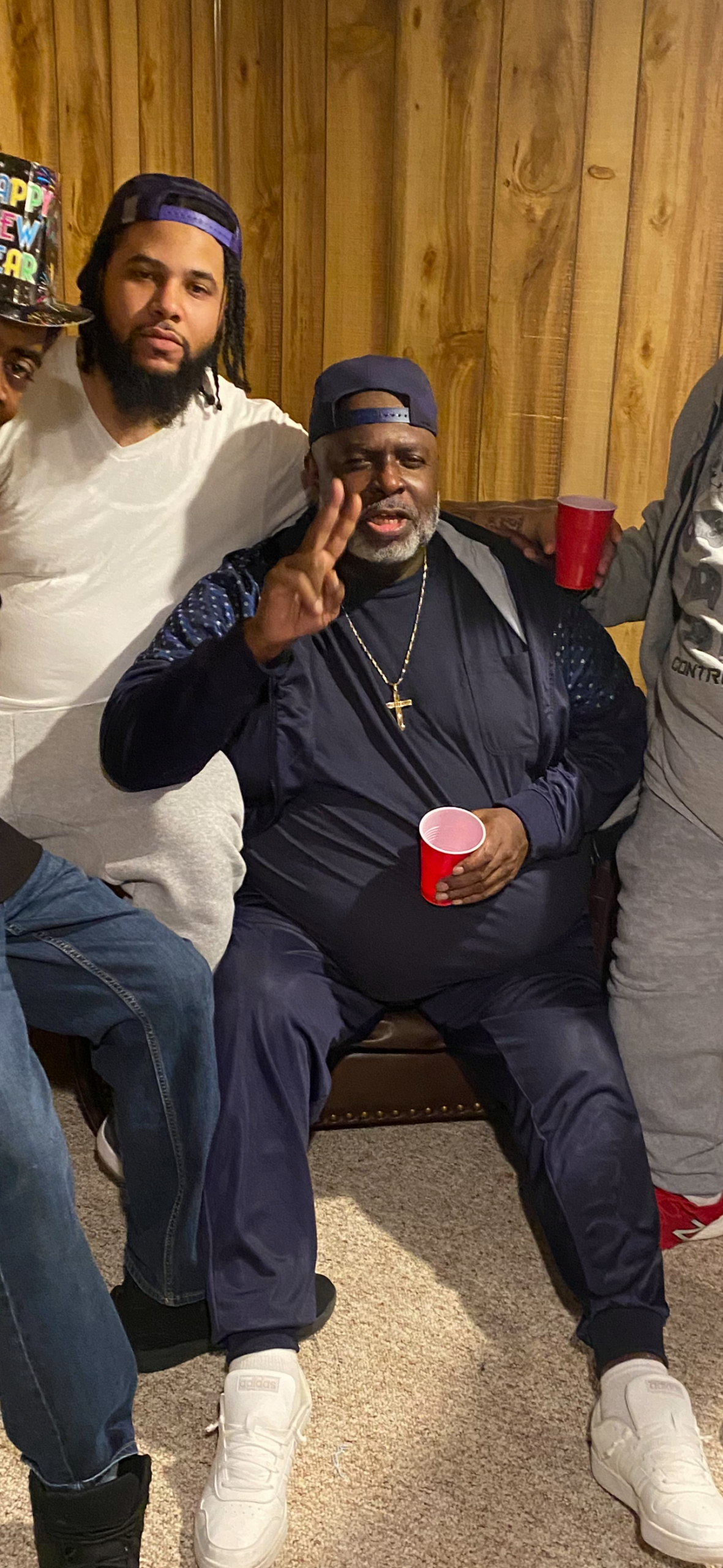 Man in a navy tracksuit seated, flashing a peace sign and wearing a cross necklace, holding a red cup; friends beside him in a wood-paneled room.