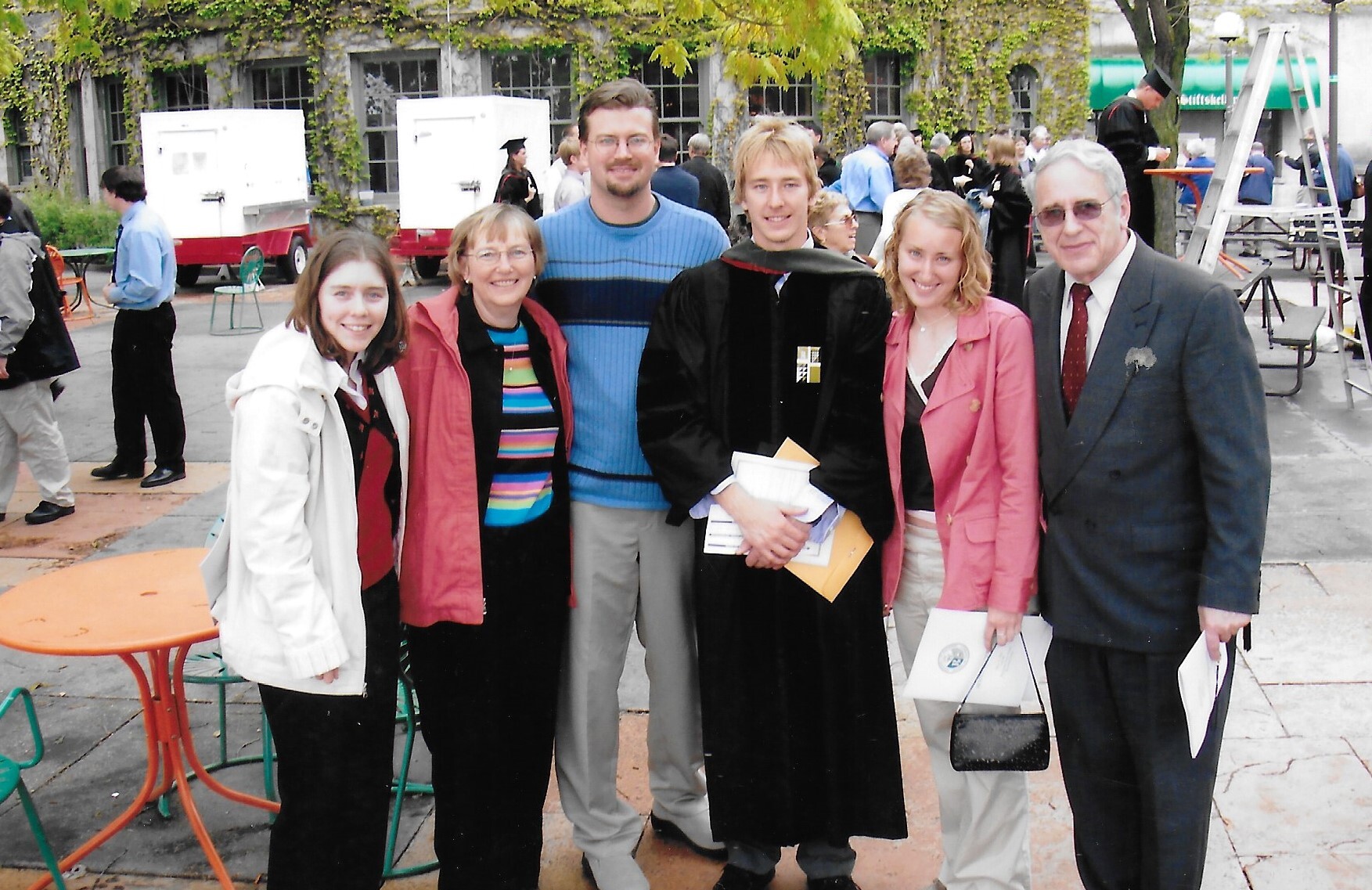 Group of six people posing for a photo at an outdoor graduation ceremony; one graduate wears a black cap and gown and holds folders and papers.