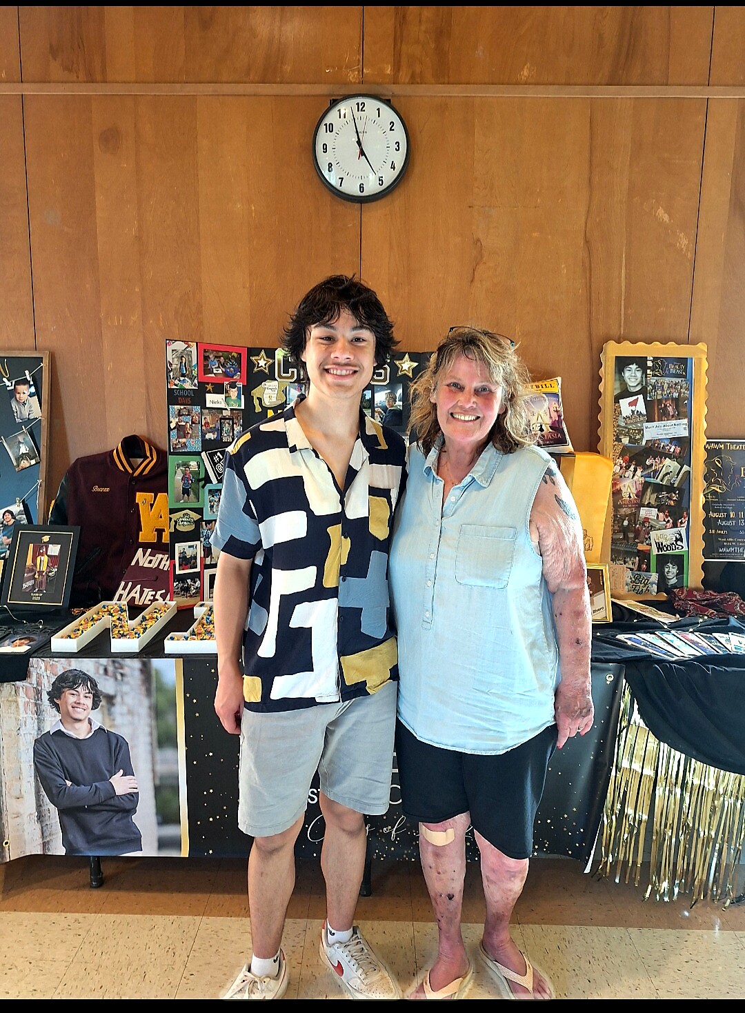 Two people pose together at a school event in front of a display board filled with photos and memorabilia.
