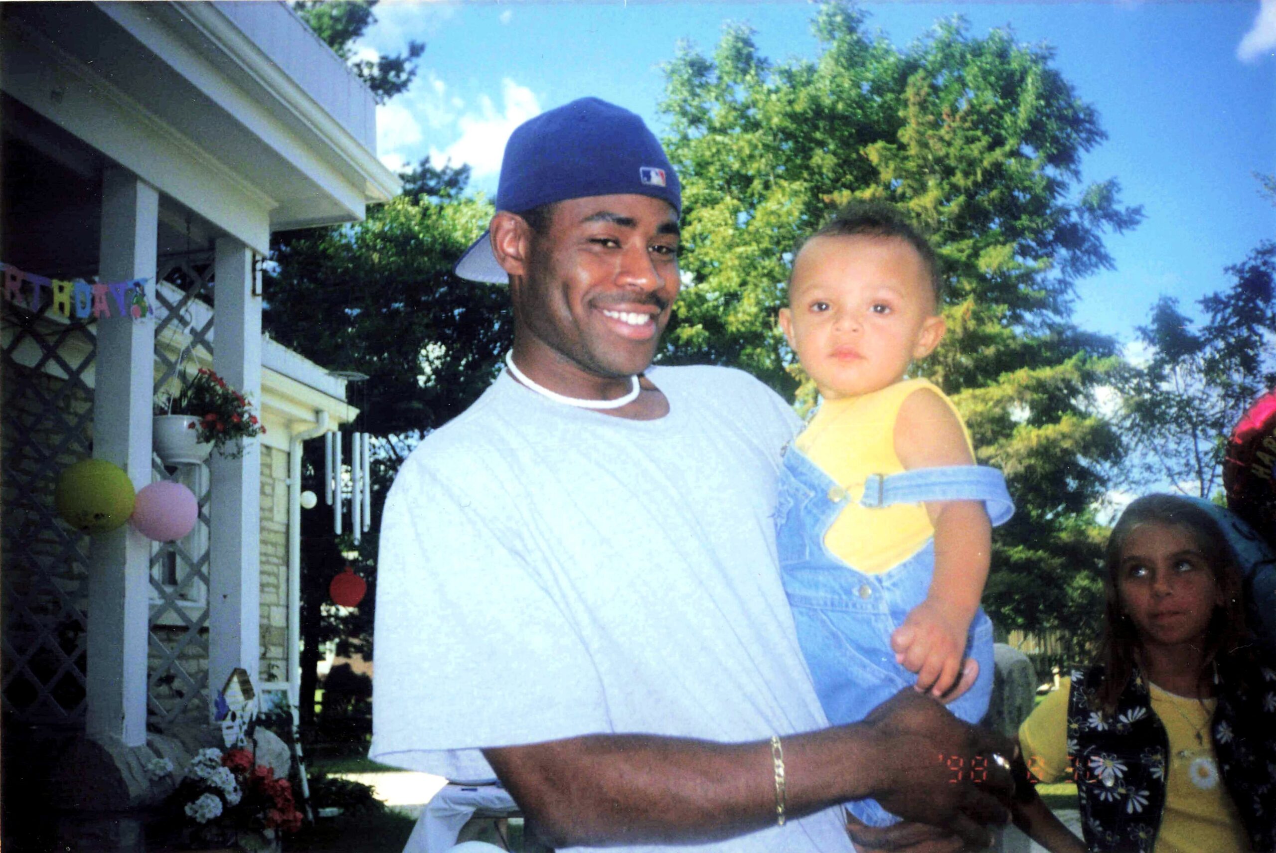 Man in a blue cap holding a young child in a yellow shirt at an outdoor birthday party, with balloons and banners in a yard.