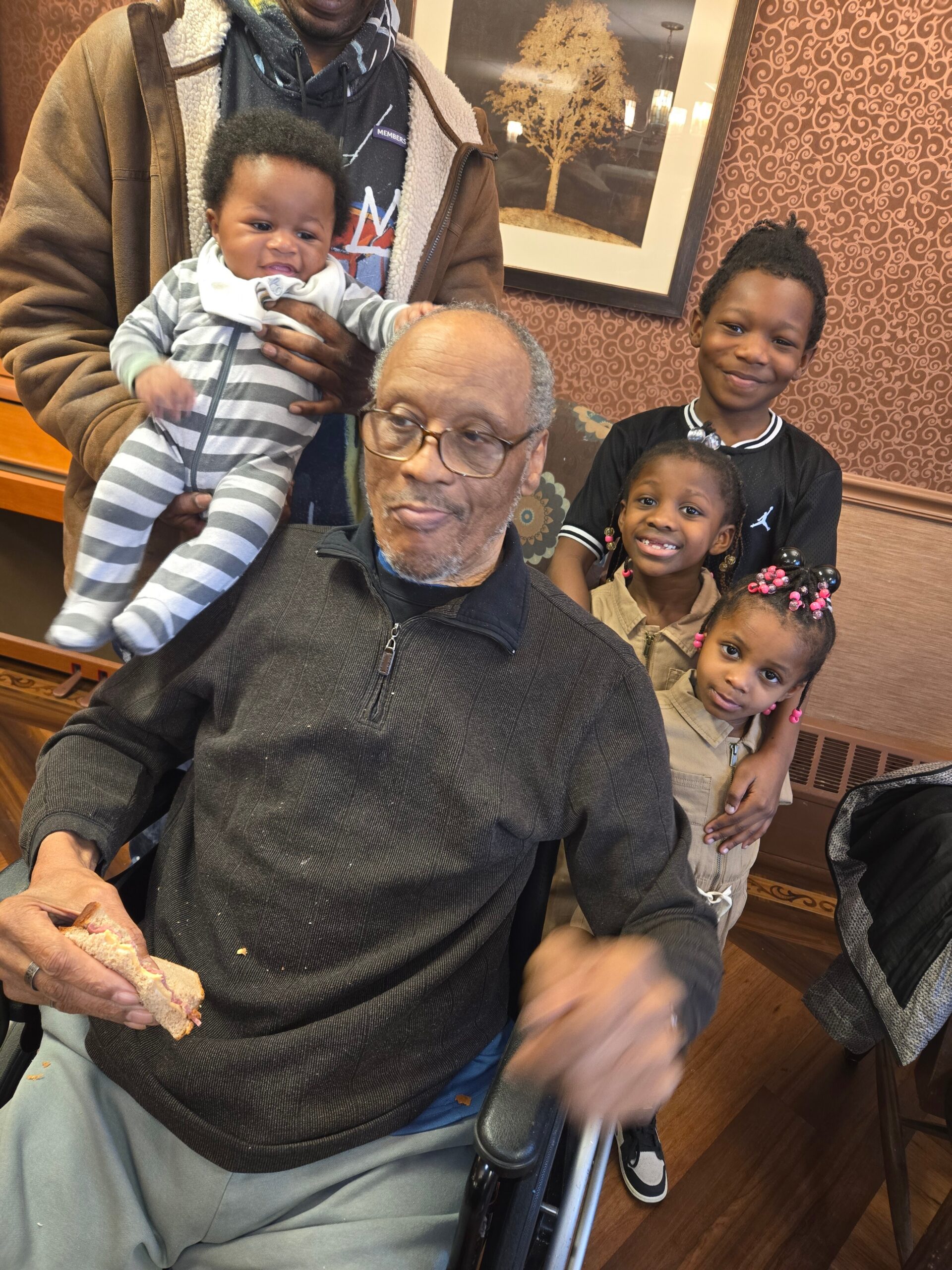 Older man in a wheelchair eating a sandwich while a man holds a baby and three kids smile nearby in a cafe.